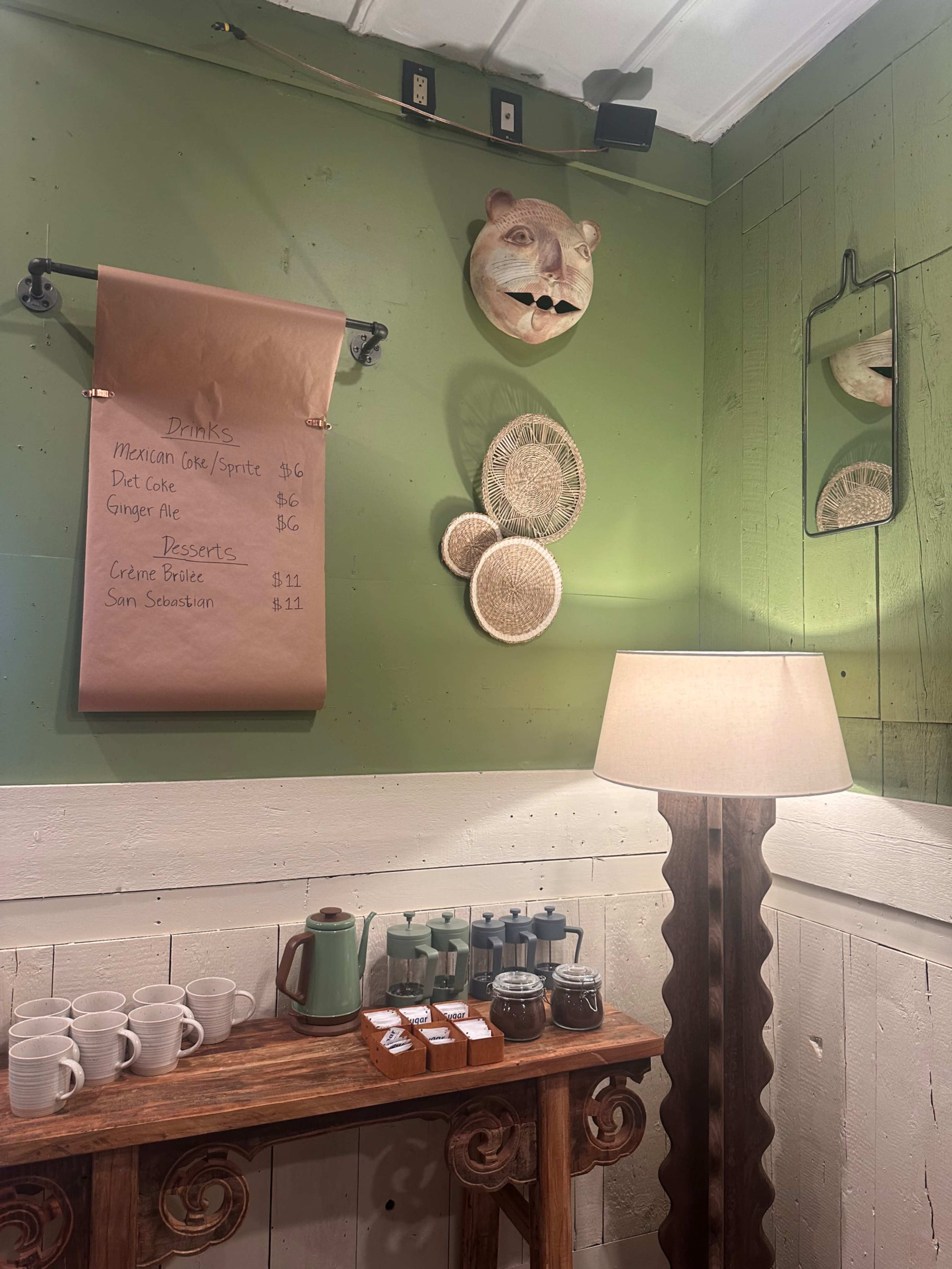 The image depicts a cozy cafe corner featuring a green wall adorned with decorative items, a menu on brown paper listing drinks and desserts, and a lamp beside a wooden table with various containers.