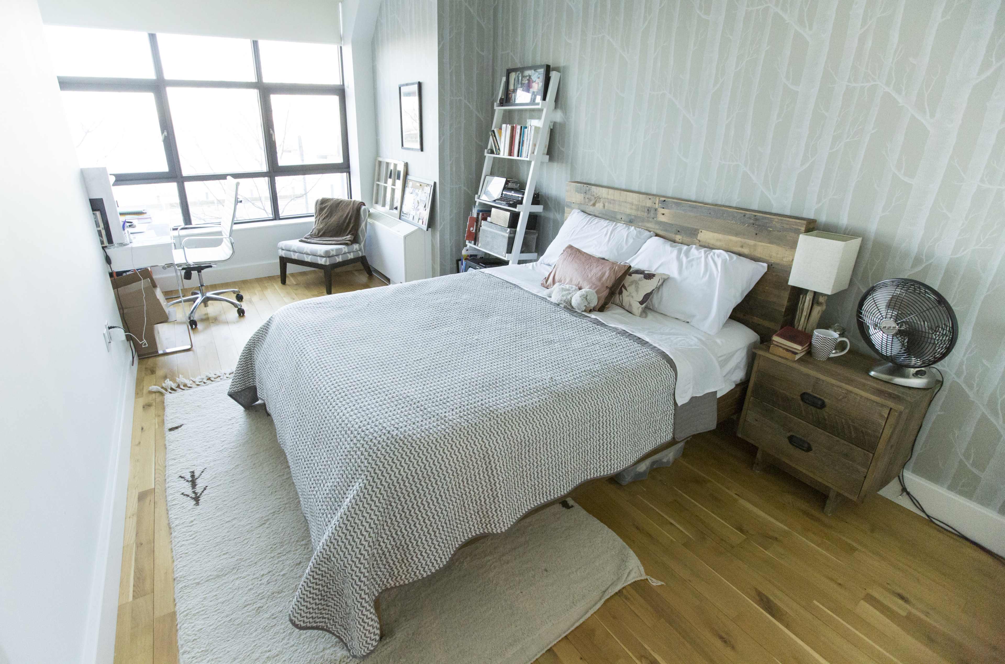 A neatly organized bedroom features a wooden bed with a light-colored blanket, a chair, a small desk, and large windows allowing natural light to enter.