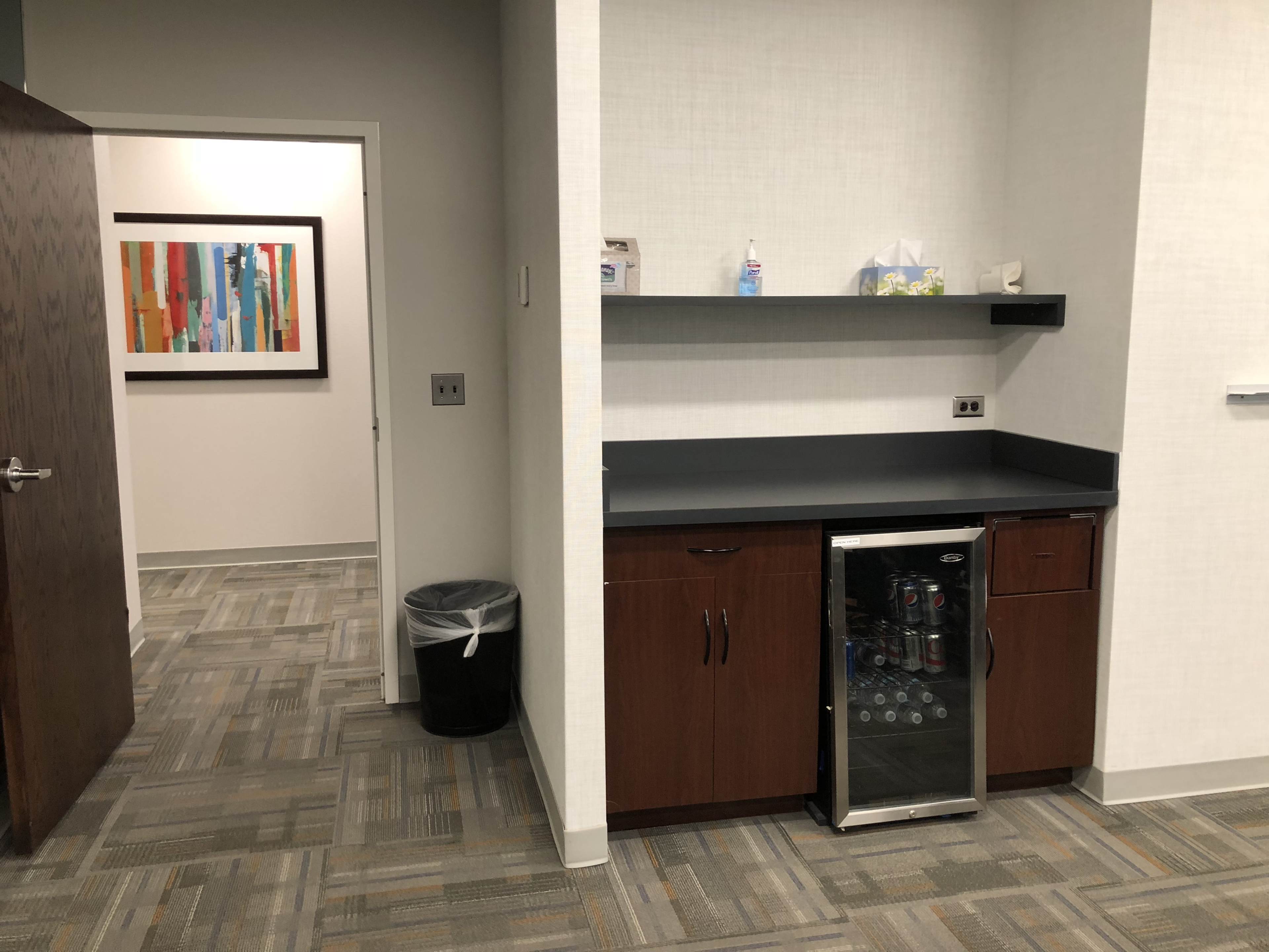 The image shows a small office kitchenette area with a black countertop, a refrigerator, and an adjacent door leading to another room.