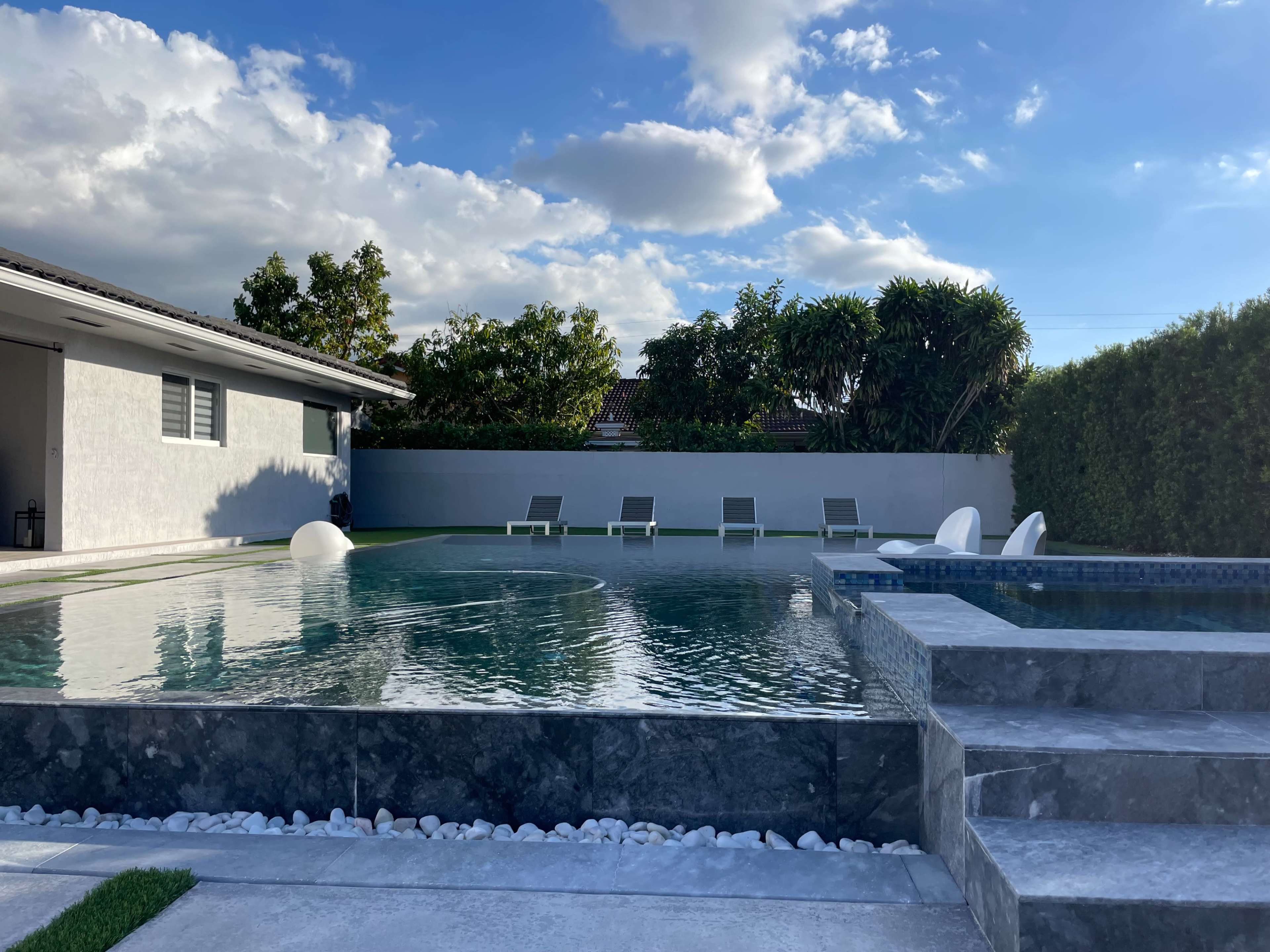 The image shows a modern swimming pool area with lounge chairs, surrounded by greenery and a house in the background under a partly cloudy sky.