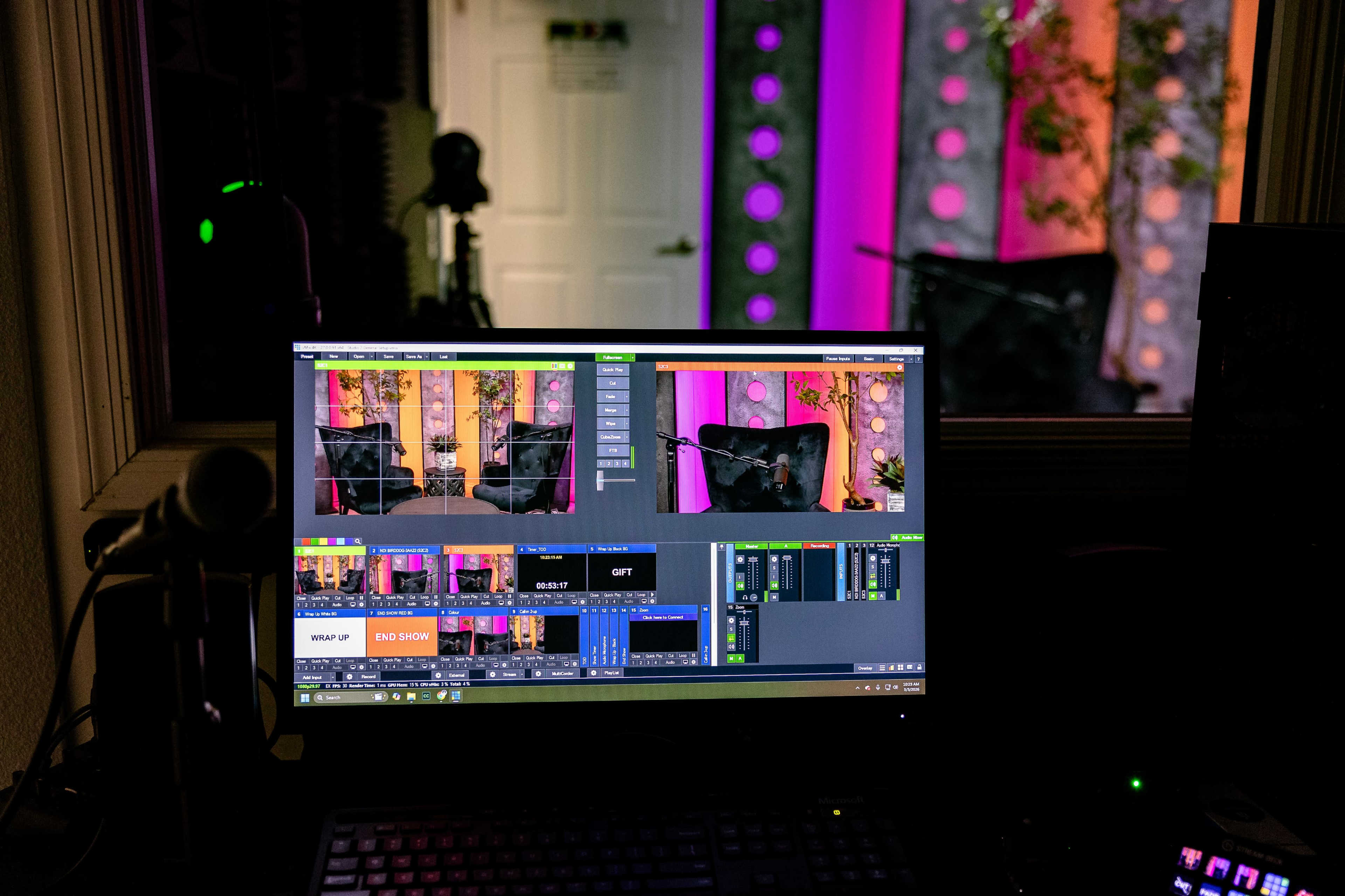 A computer monitor displays multiple camera feeds and production controls in a studio environment with colorful lighting.