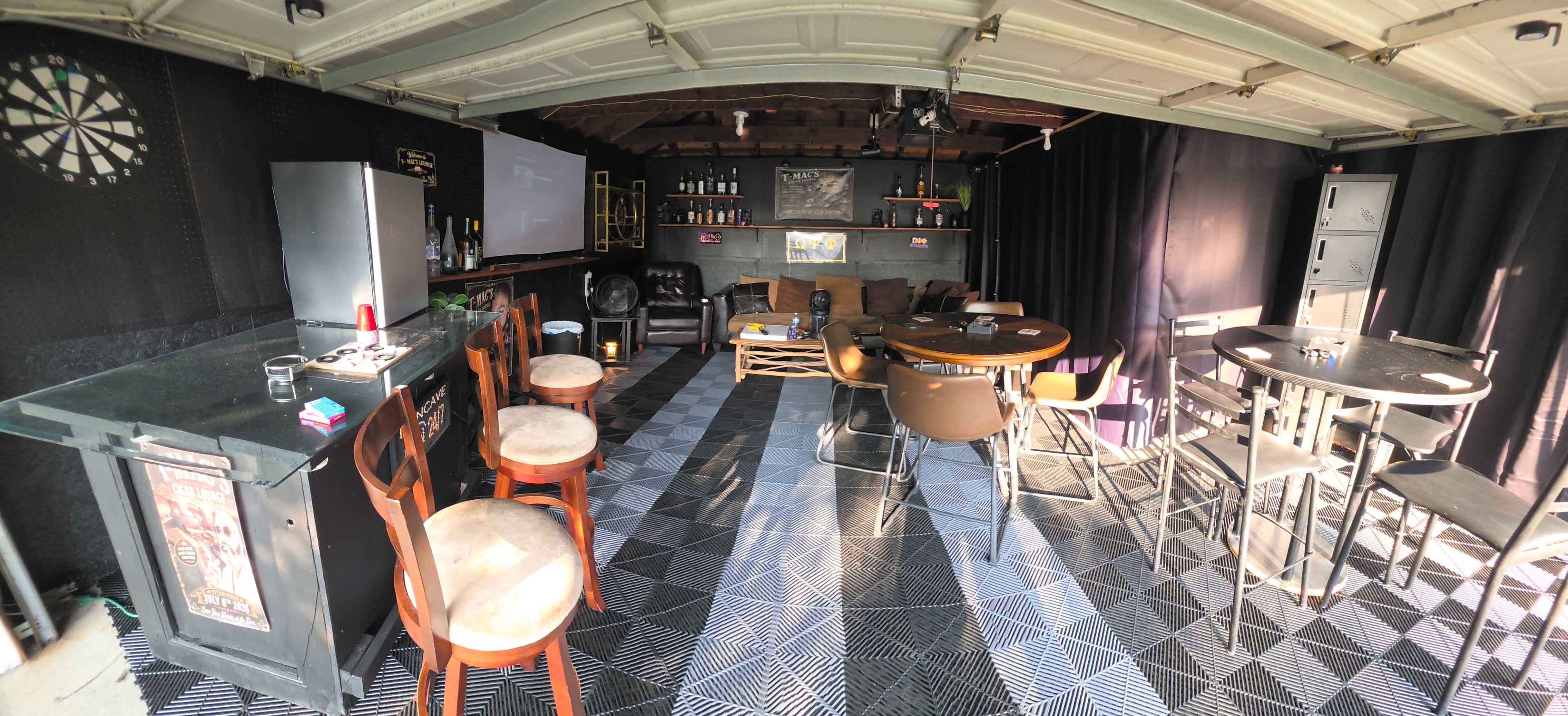 The image depicts a home bar setup inside a garage, featuring seating areas, a dartboard, a bar counter, and shelves filled with bottles.