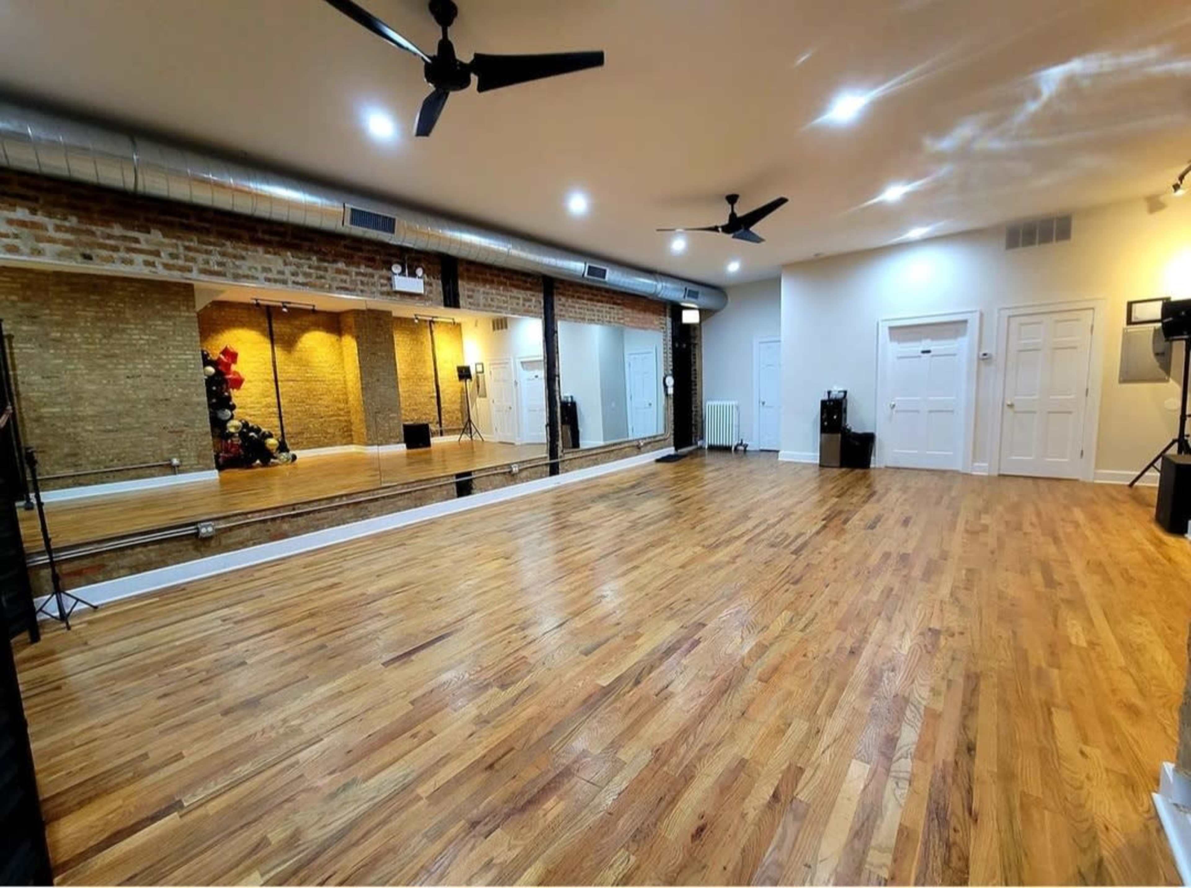 The image shows a spacious dance studio with polished wooden floors, mirrors on one wall, and large ceiling fans.