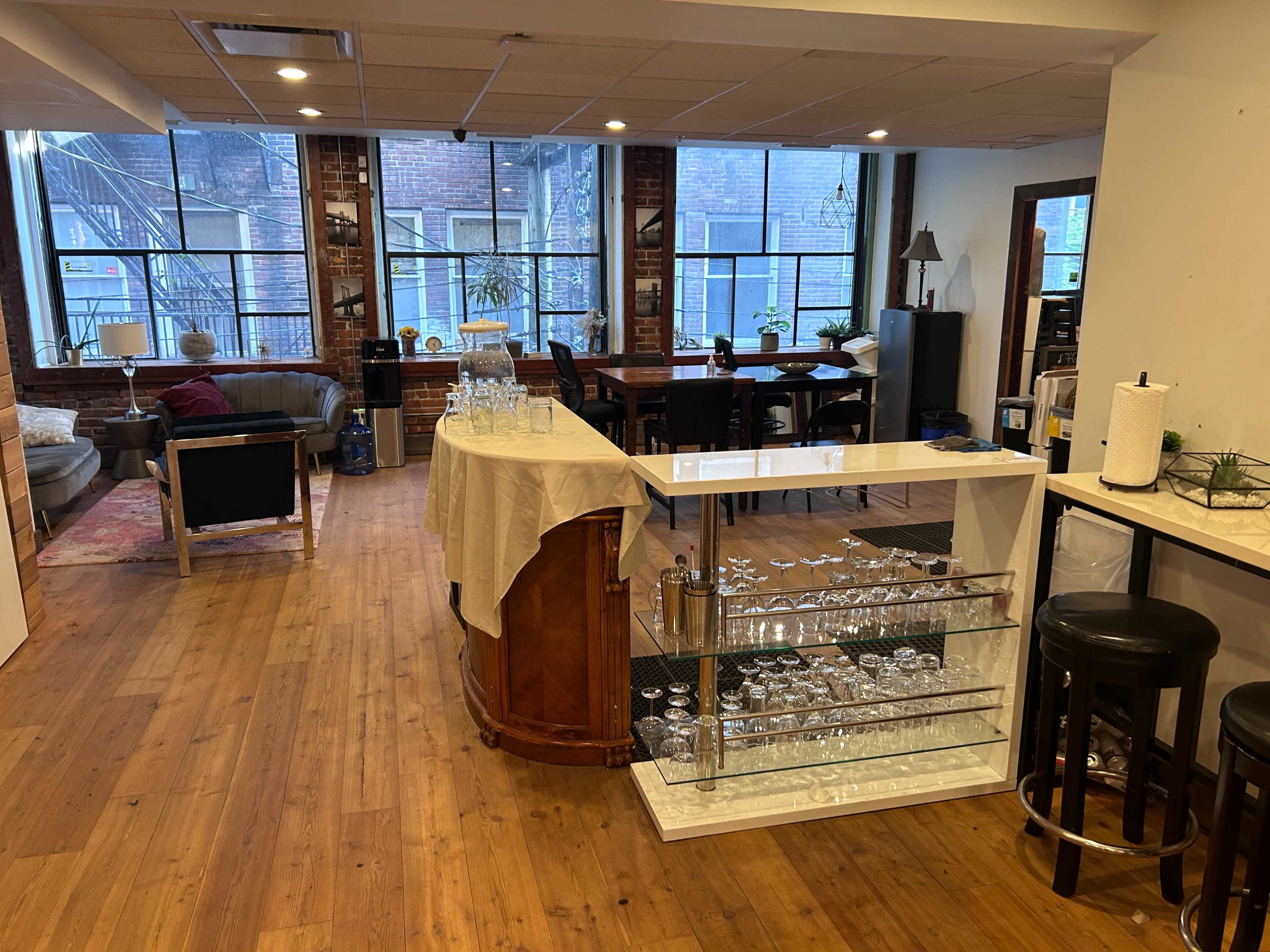 The image shows a spacious, well-lit room with wooden flooring, featuring a bar area with glassware, seating arrangements including sofas and tables, and large windows providing natural light.