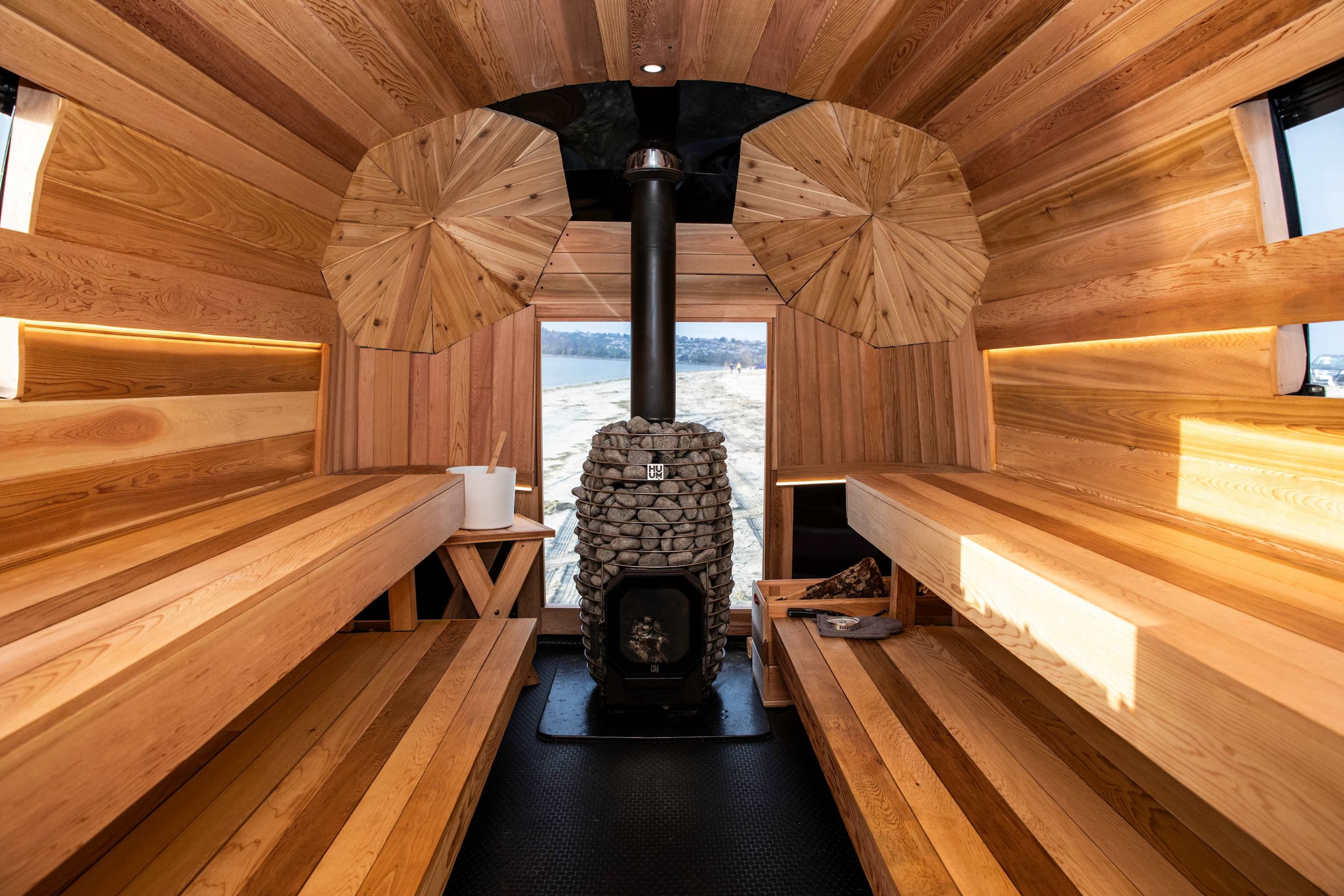 The interior of a wooden sauna features two wooden benches on either side, a stone wood stove in the center, and large windows allowing natural light to enter.