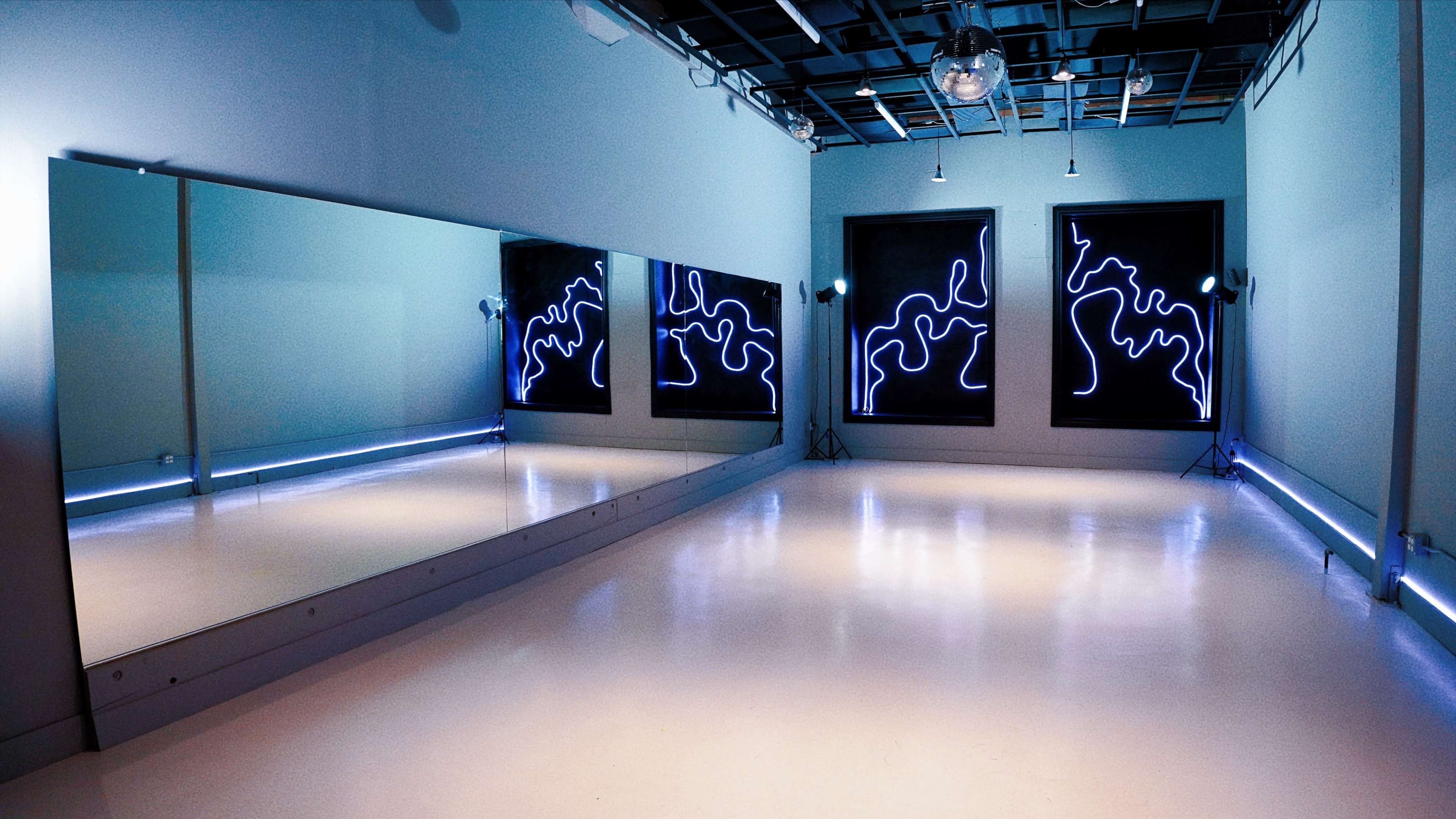 A dance studio featuring large mirrors on one wall, abstract illuminated art on the opposite wall, and a smooth white floor.