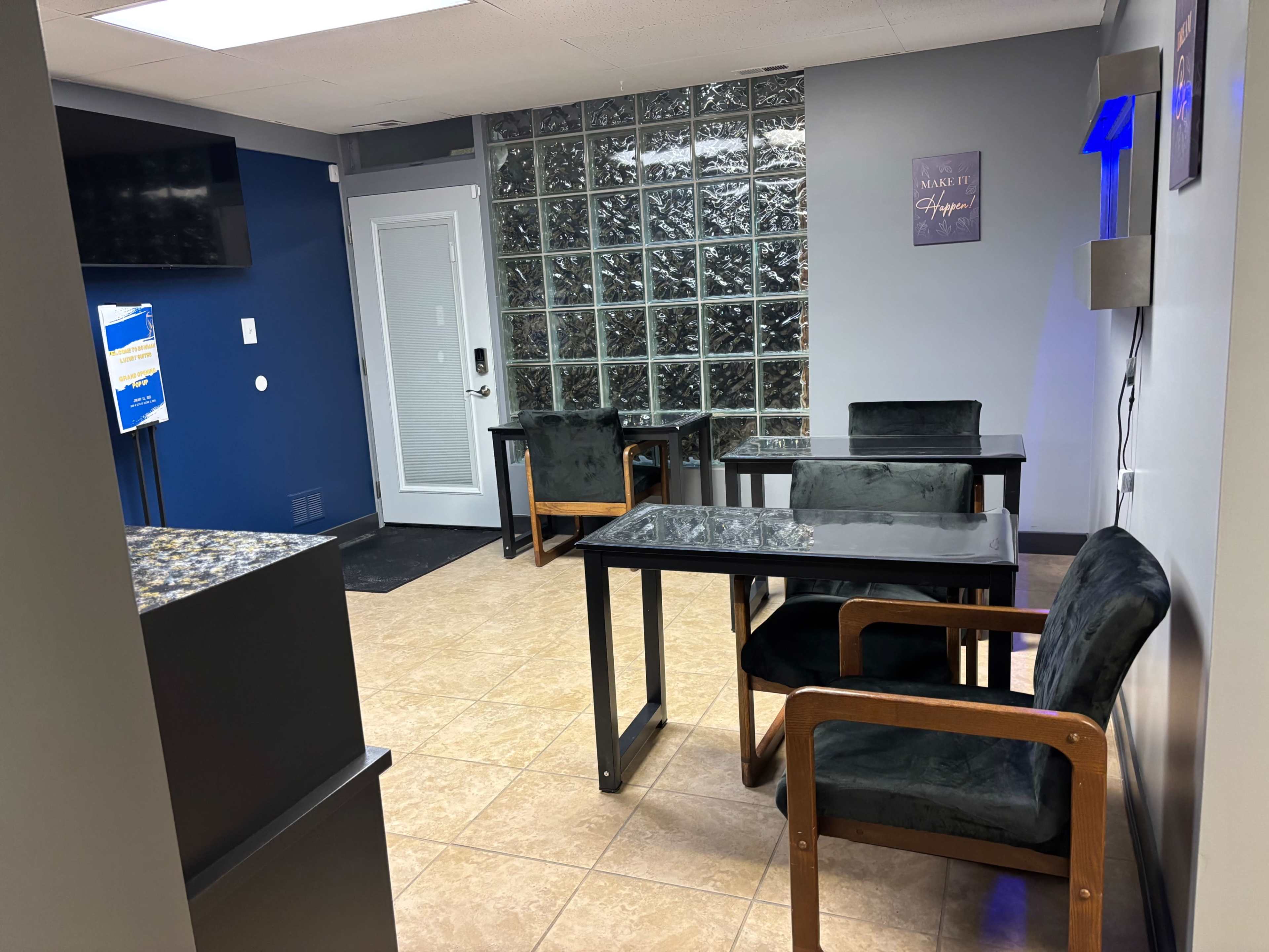 The image shows a small office waiting area with glass block walls, two black chairs, two black tables, and a door leading outside.