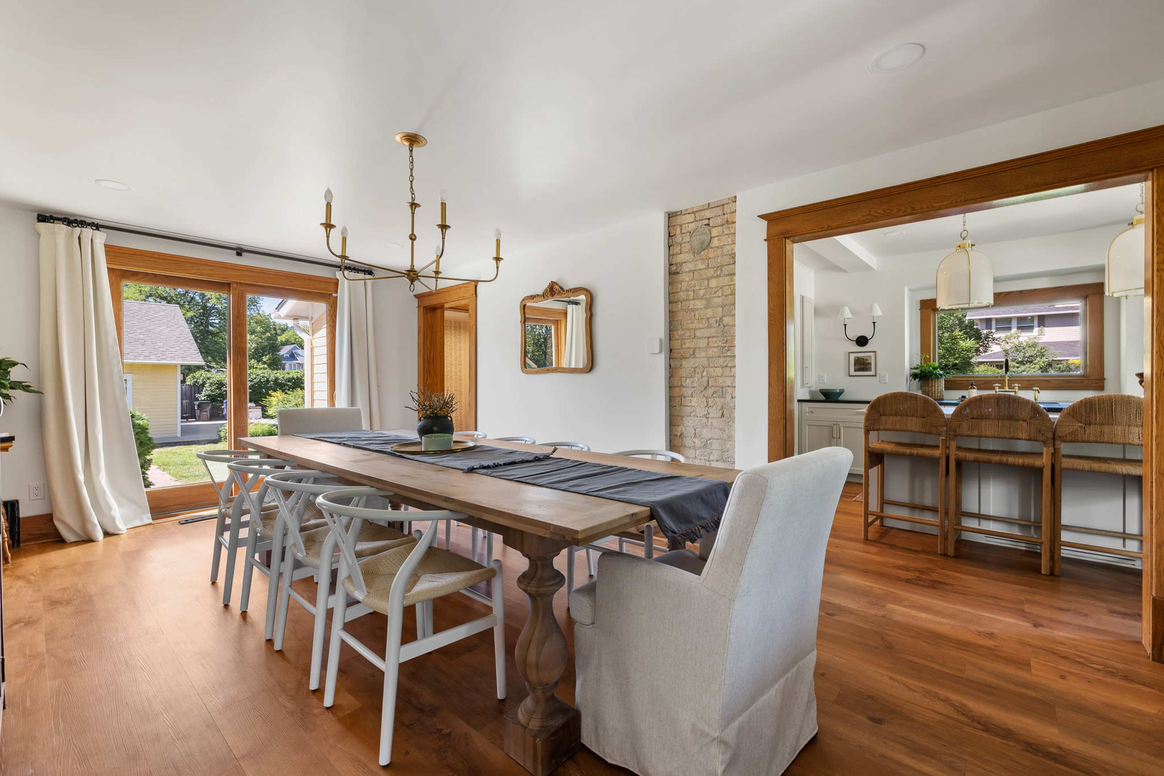 A dining room with a large wooden table surrounded by white chairs, featuring a chandelier and an open view to an outdoor area through large glass doors.