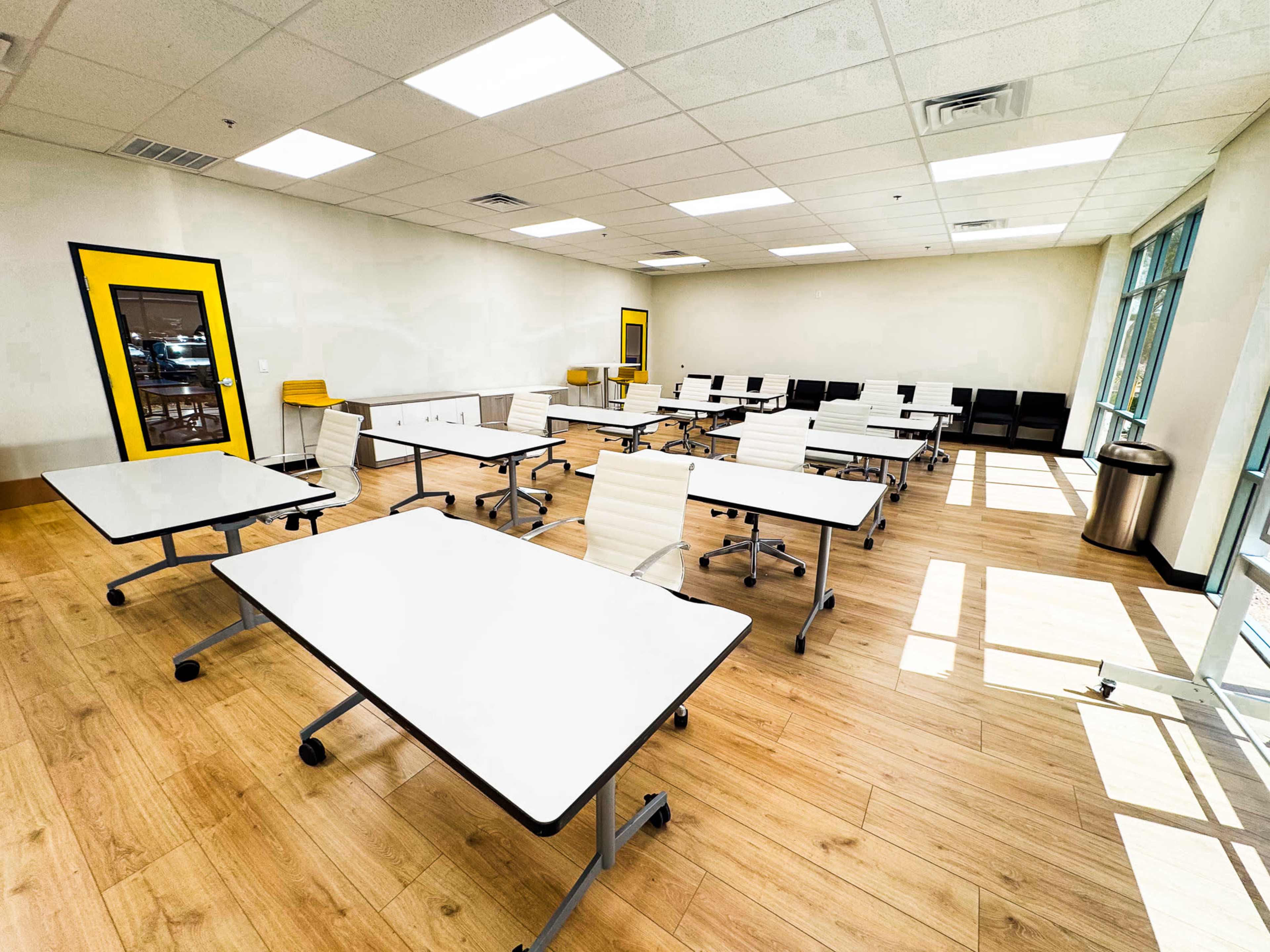 The image shows a spacious classroom with several white tables and chairs arranged in rows, large windows allowing natural light, and a yellow-framed door.