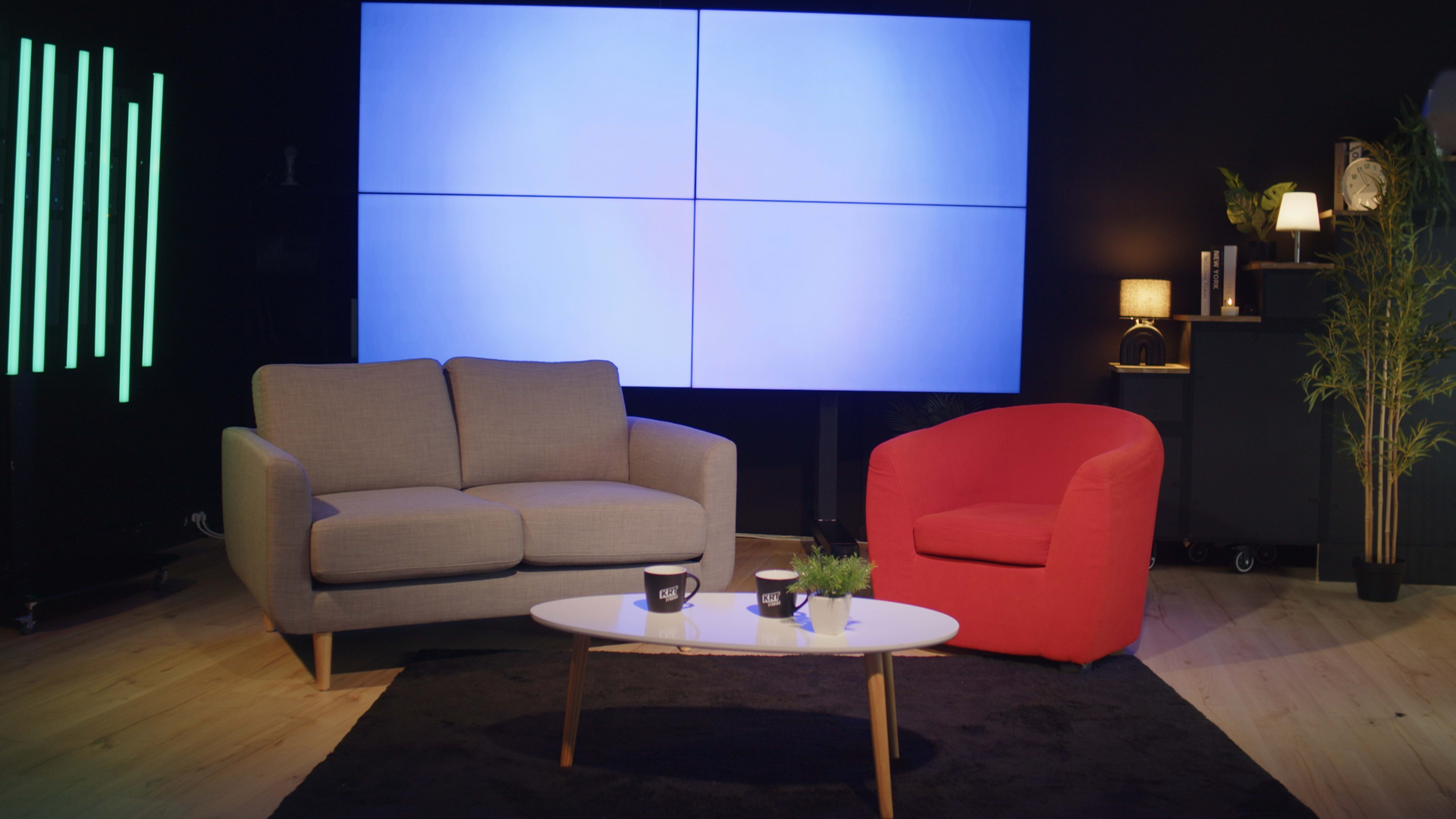 The image shows a modern studio setup featuring a gray sofa and a red armchair positioned around a white coffee table on a black rug, with a large screen display in the background.