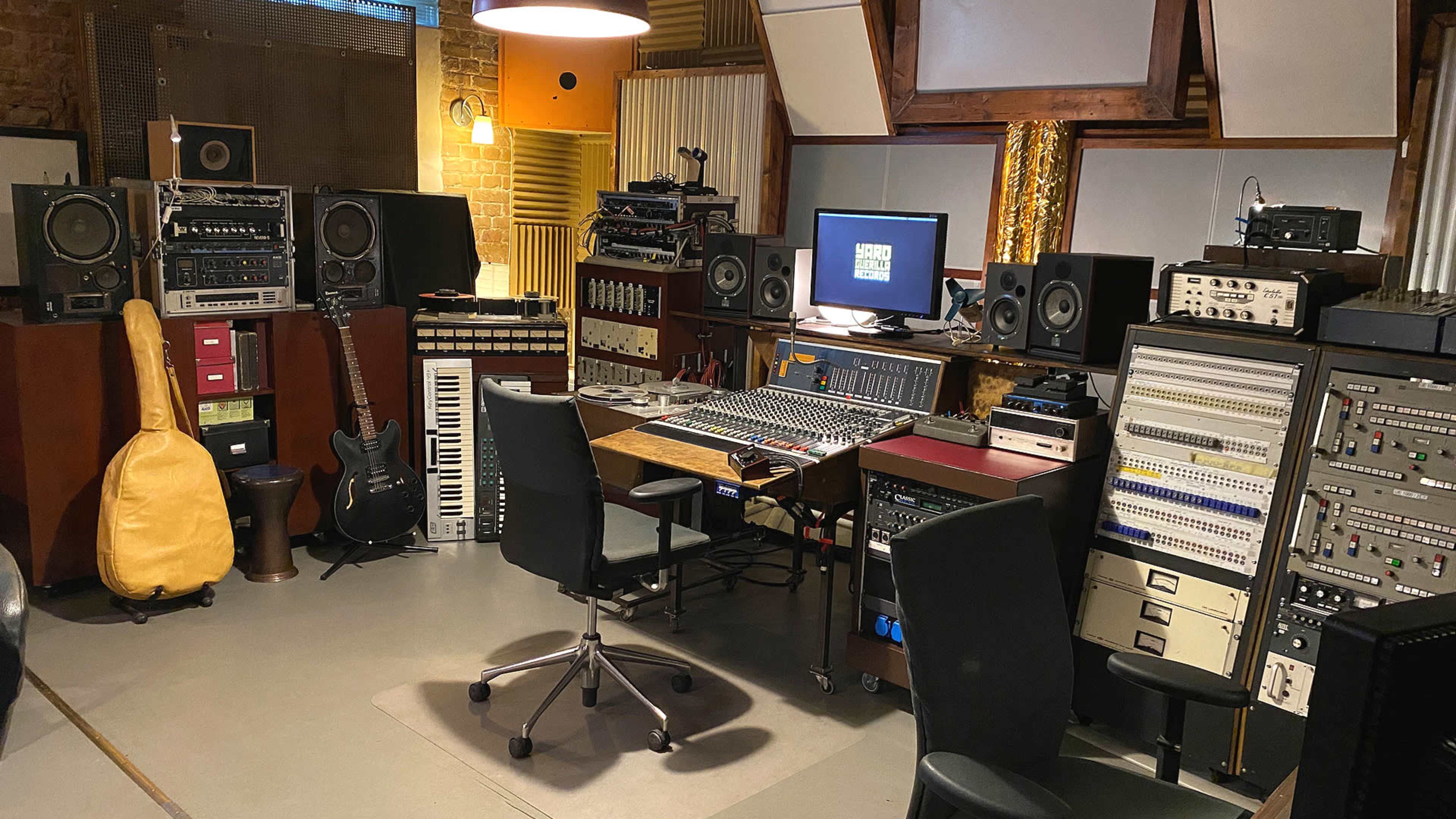 A music recording studio features a central mixing console surrounded by various audio equipment, instruments, and monitors.