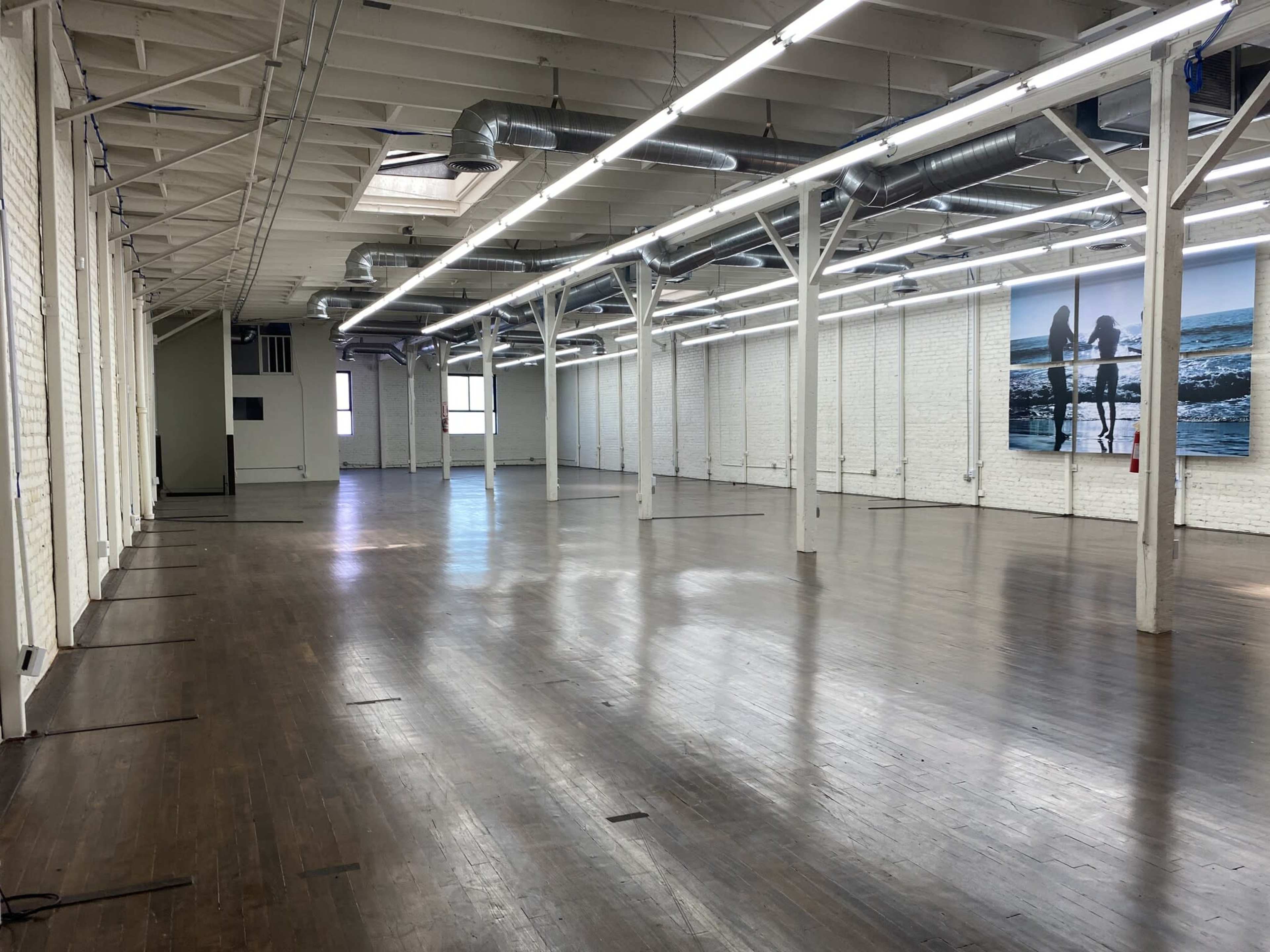 A spacious, empty loft with wooden floors and exposed beams, featuring white brick walls and large windows.