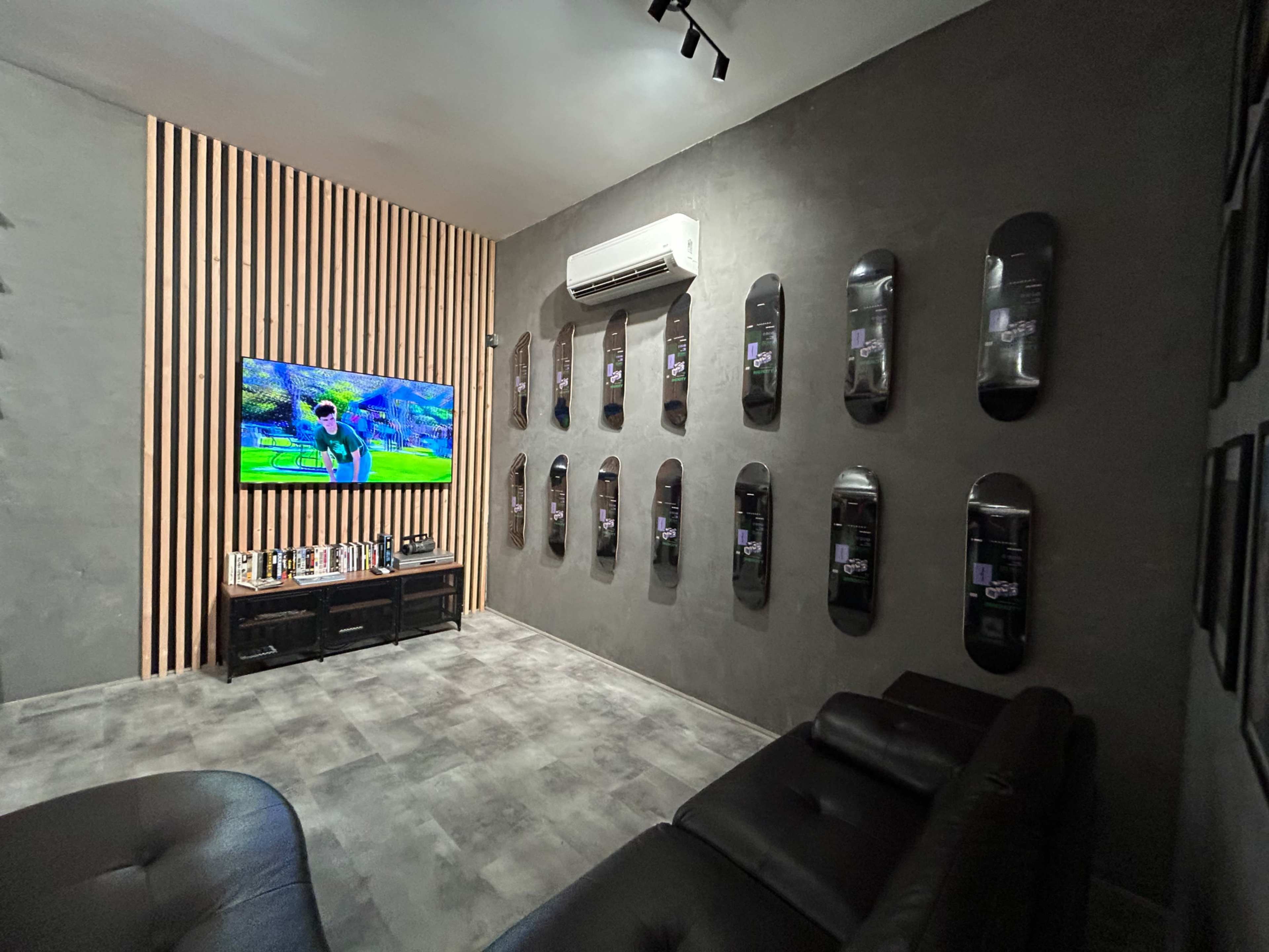 The room features a television displaying a video game, surrounded by skateboard decks arranged on the wall and black leather seating.