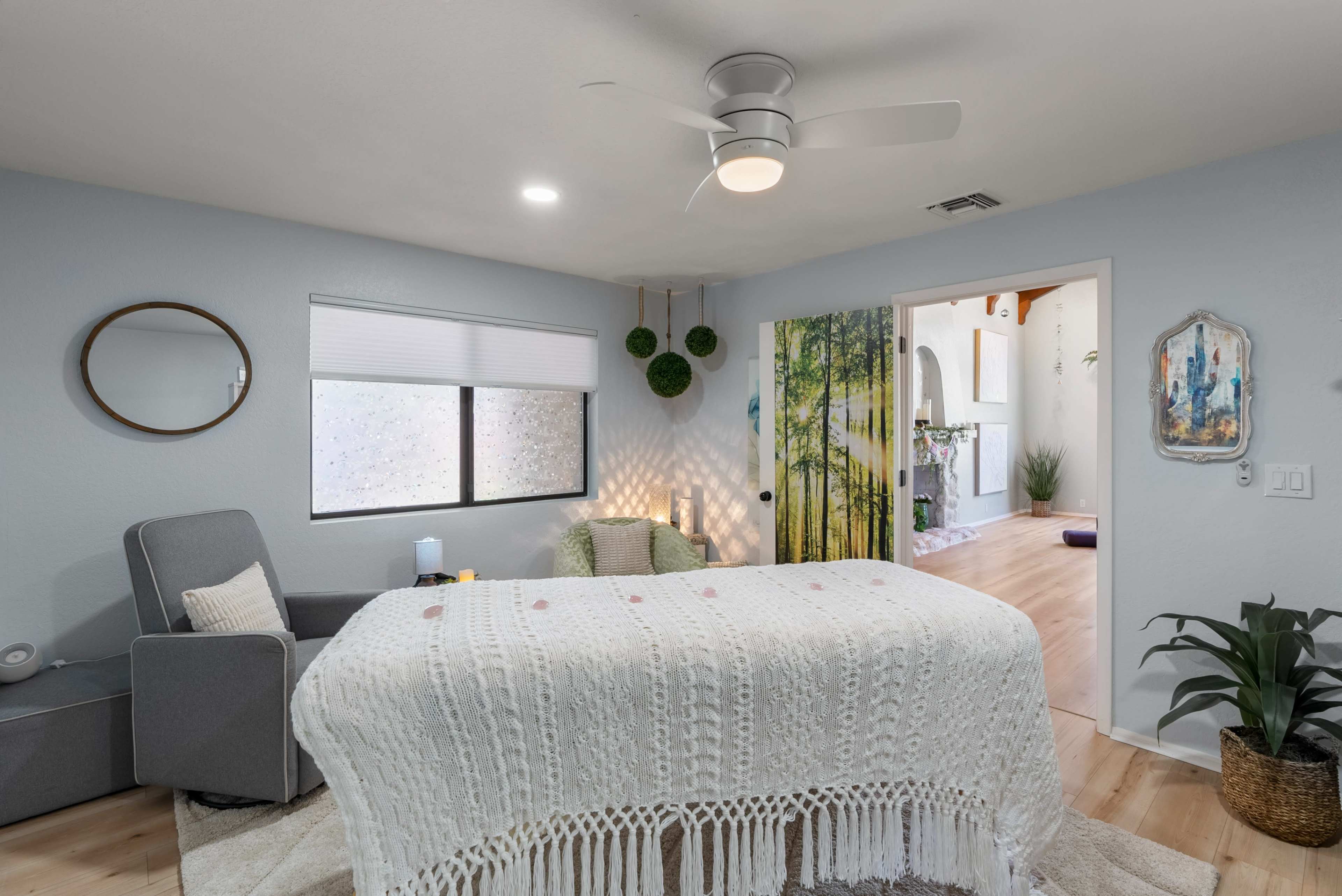 A cozy room features a bed with a white knitted throw, a gray armchair, and a door leading to another space decorated with greenery.