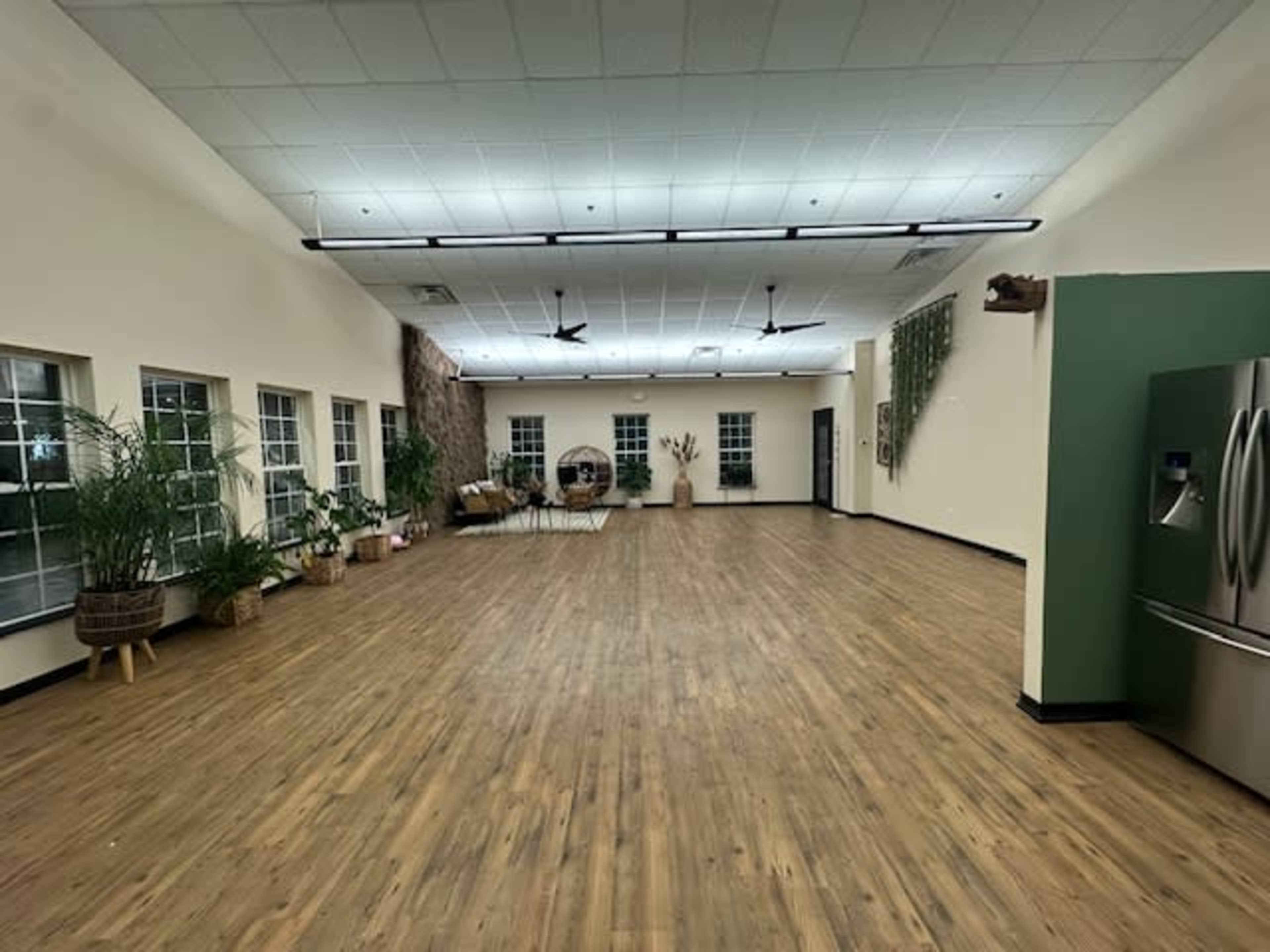 A spacious, well-lit room with wooden flooring, large windows, and various plants arranged along the walls.