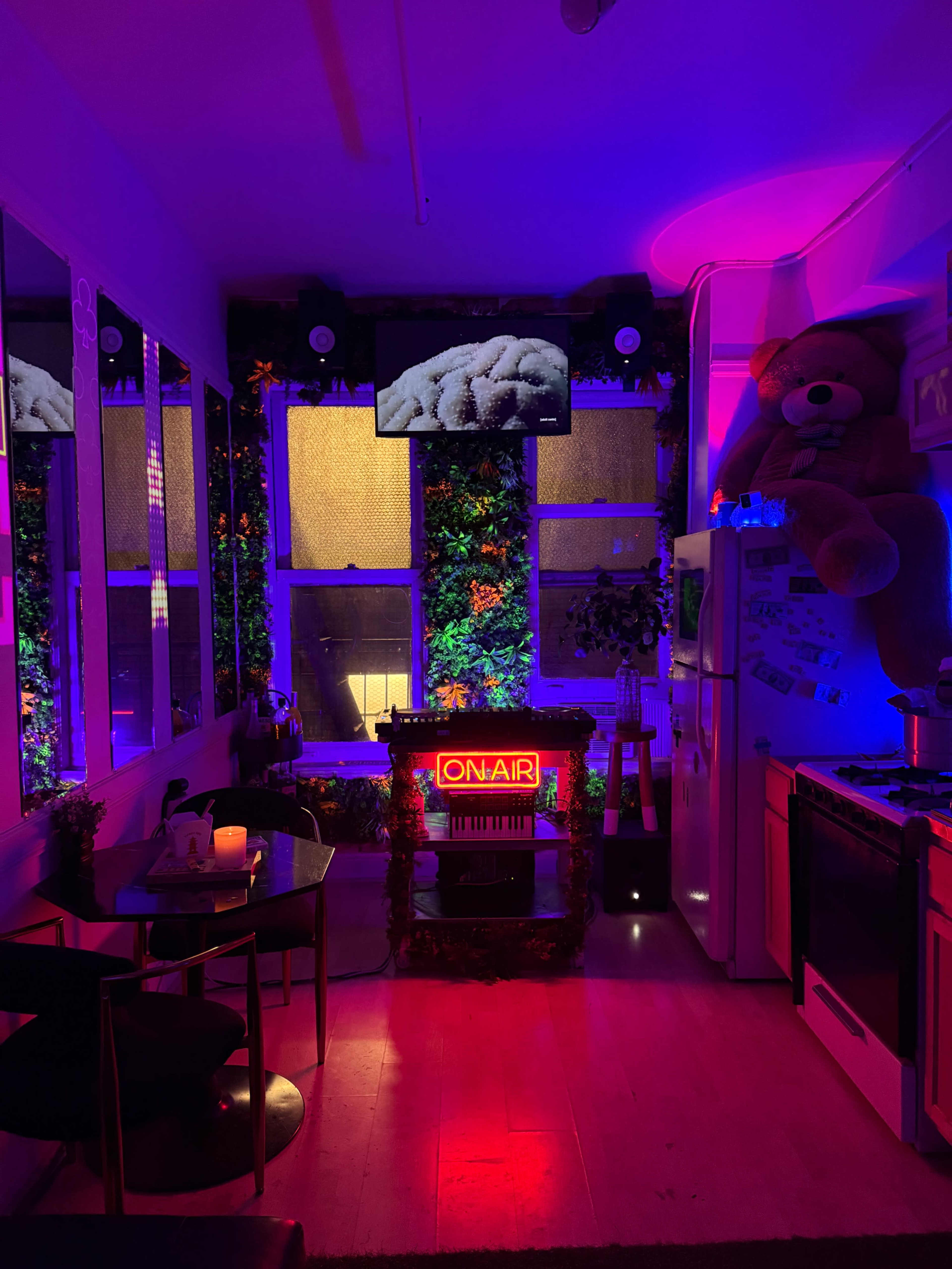 The image showcases a small kitchen space adorned with colorful lighting, a large teddy bear, a neon "ON AIR" sign, and mirrors reflecting the vibrant decor.