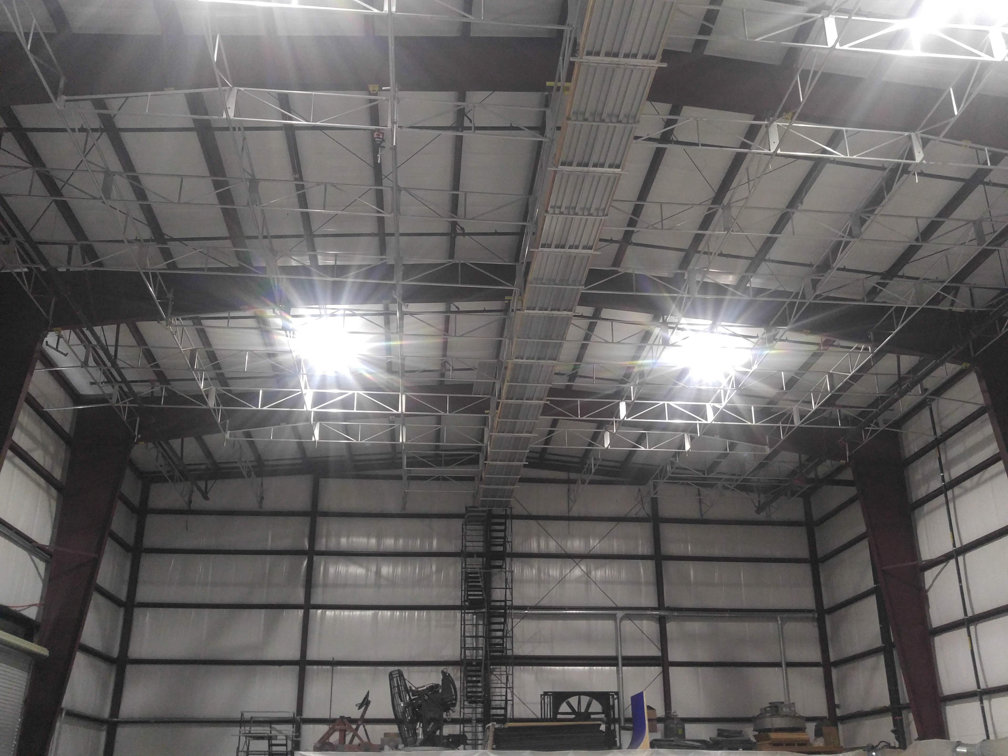 The image shows the interior of a large industrial building with a high ceiling, exposed metal beams, and bright overhead lights.