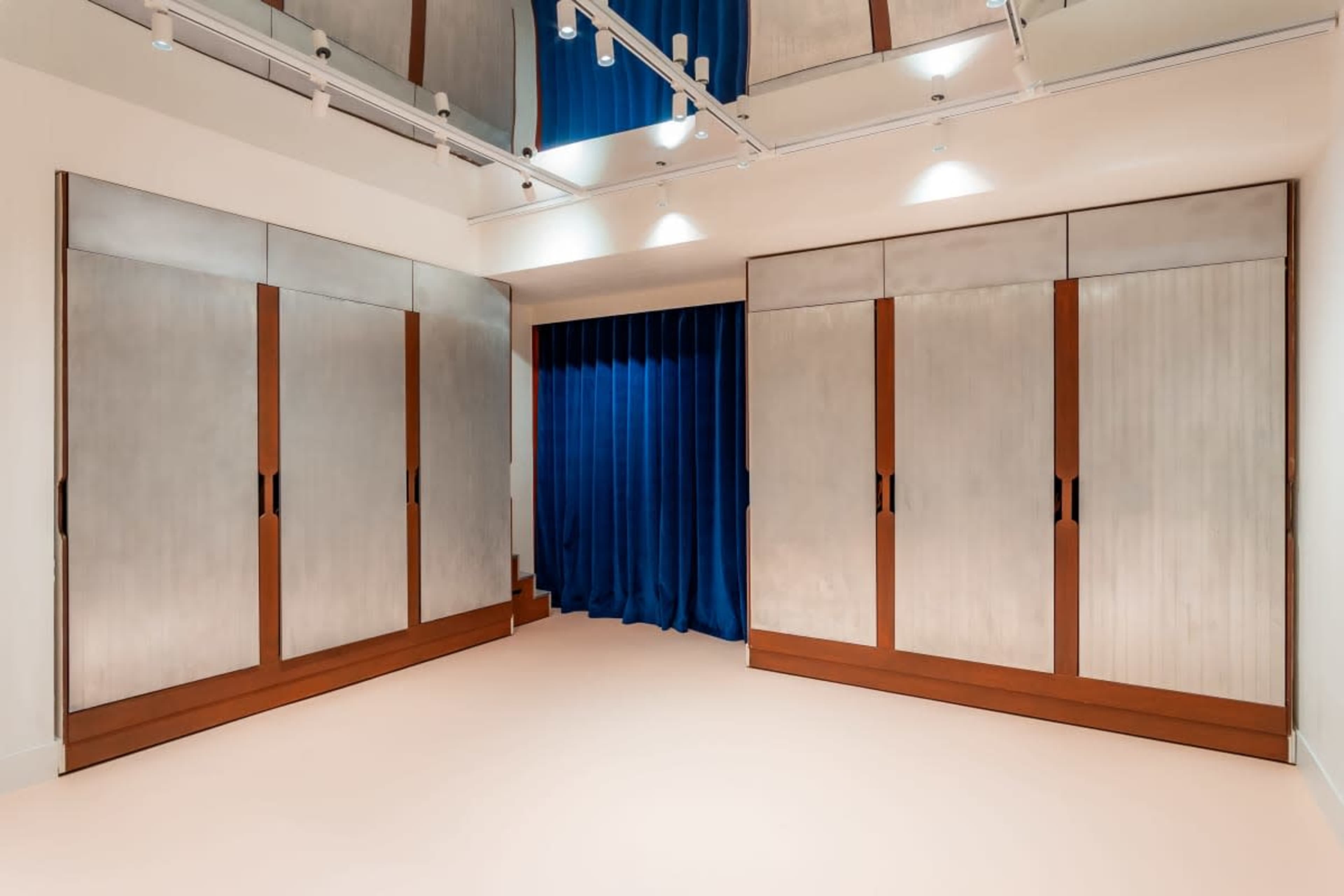 The image shows a modern room with sleek metallic cabinetry and a deep blue curtain, set against a reflective ceiling and a light-colored floor.