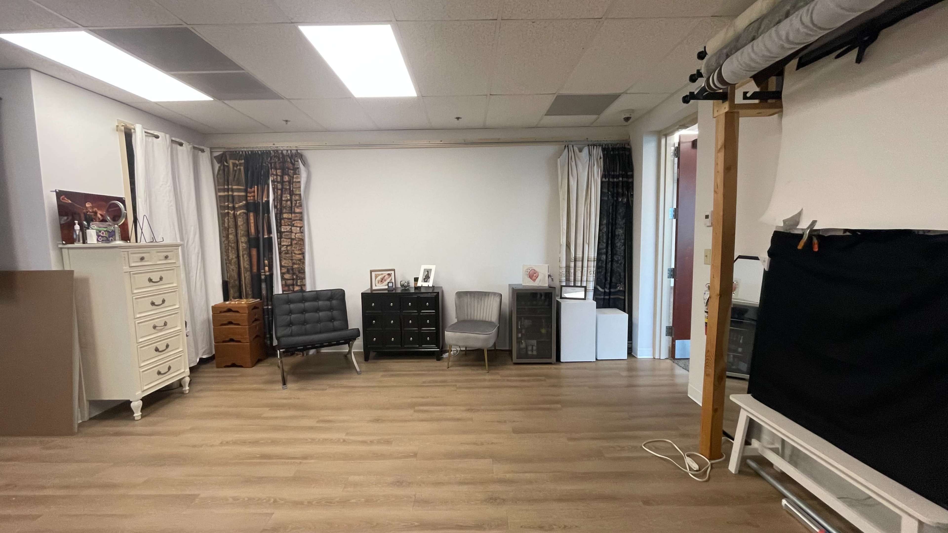 The image shows a room with wooden flooring, featuring various furniture items, including chairs, dressers, and cabinets, against white walls and curtains.