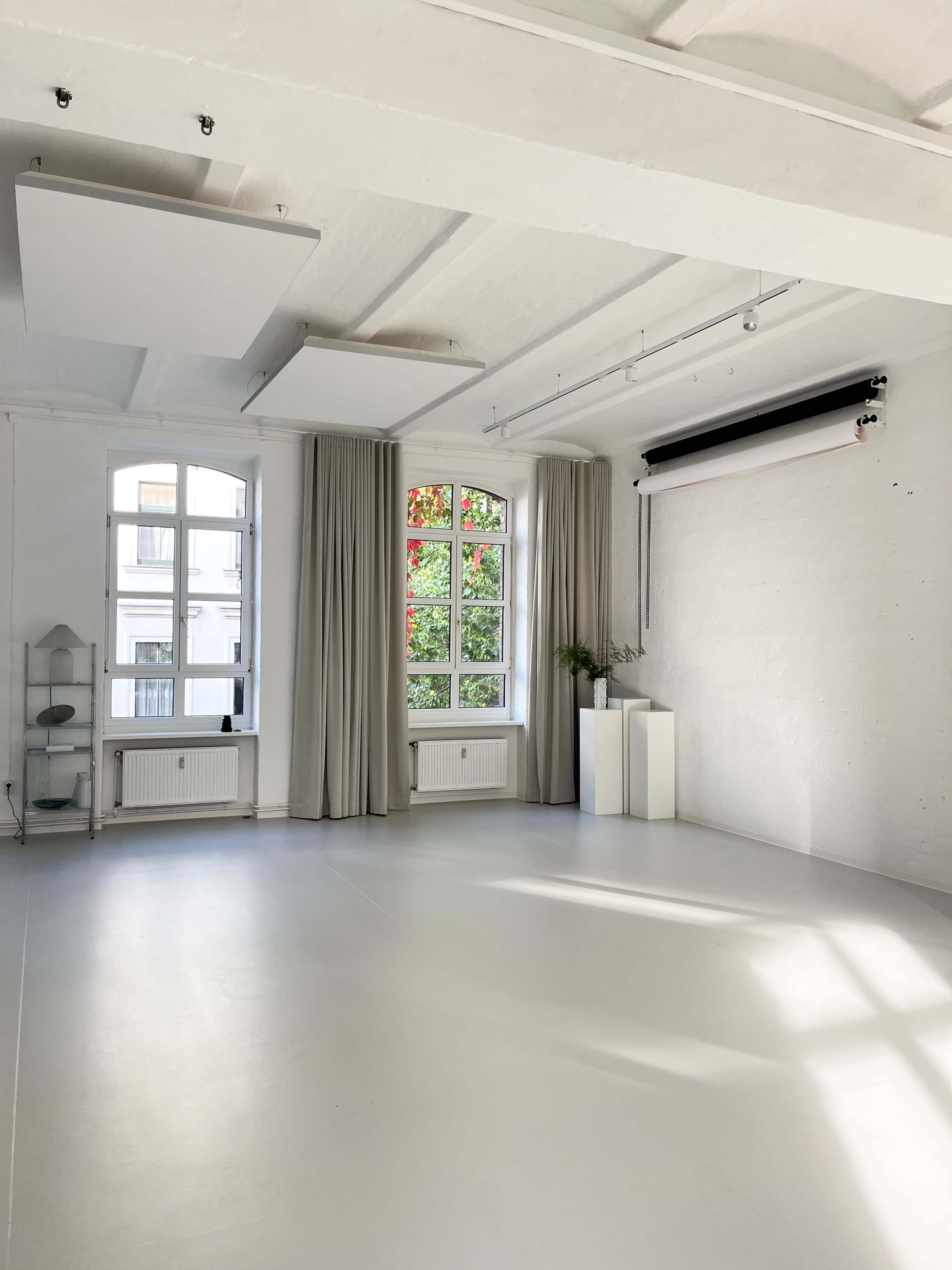 The image shows a bright, minimalist studio with large windows, white walls, and a smooth, light-colored floor.