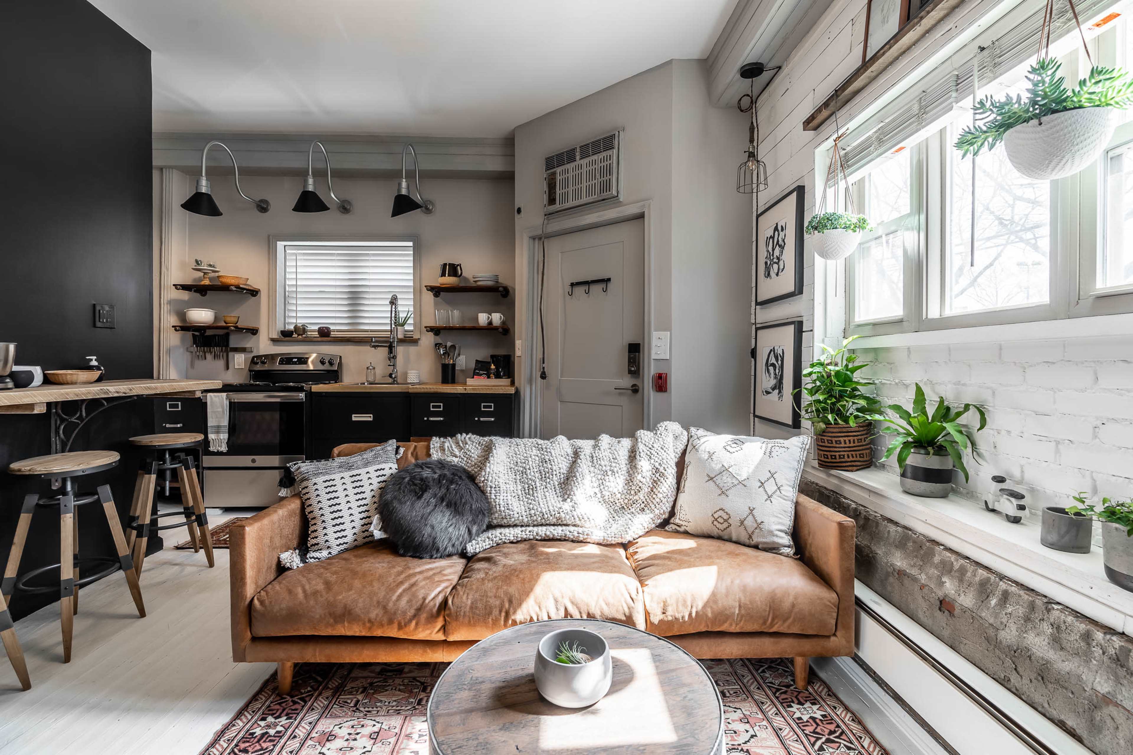 A cozy living room features a leather couch, a round table, and large windows with plants, alongside a kitchen area with dark cabinets and pendant lighting.