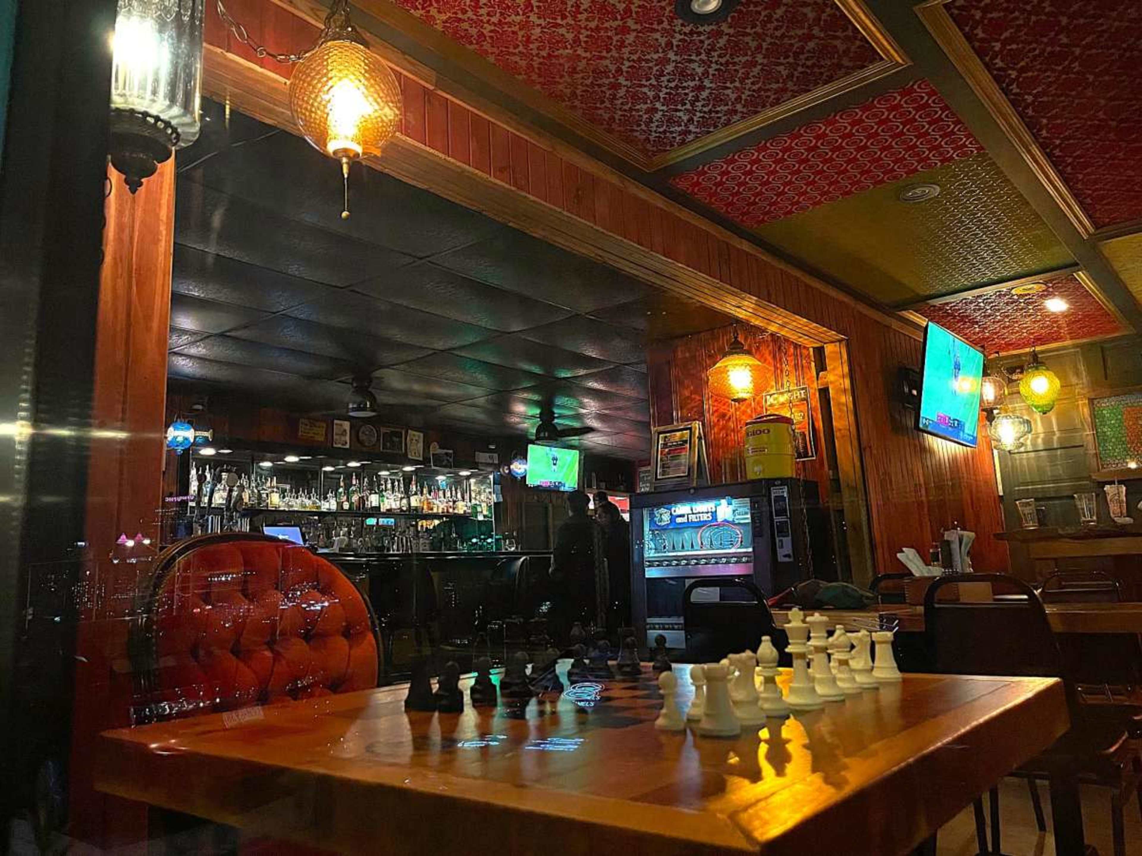 A chess set is positioned on a wooden table in a dimly lit bar, with visible bar stools and a large mirror reflecting the bar area in the background.