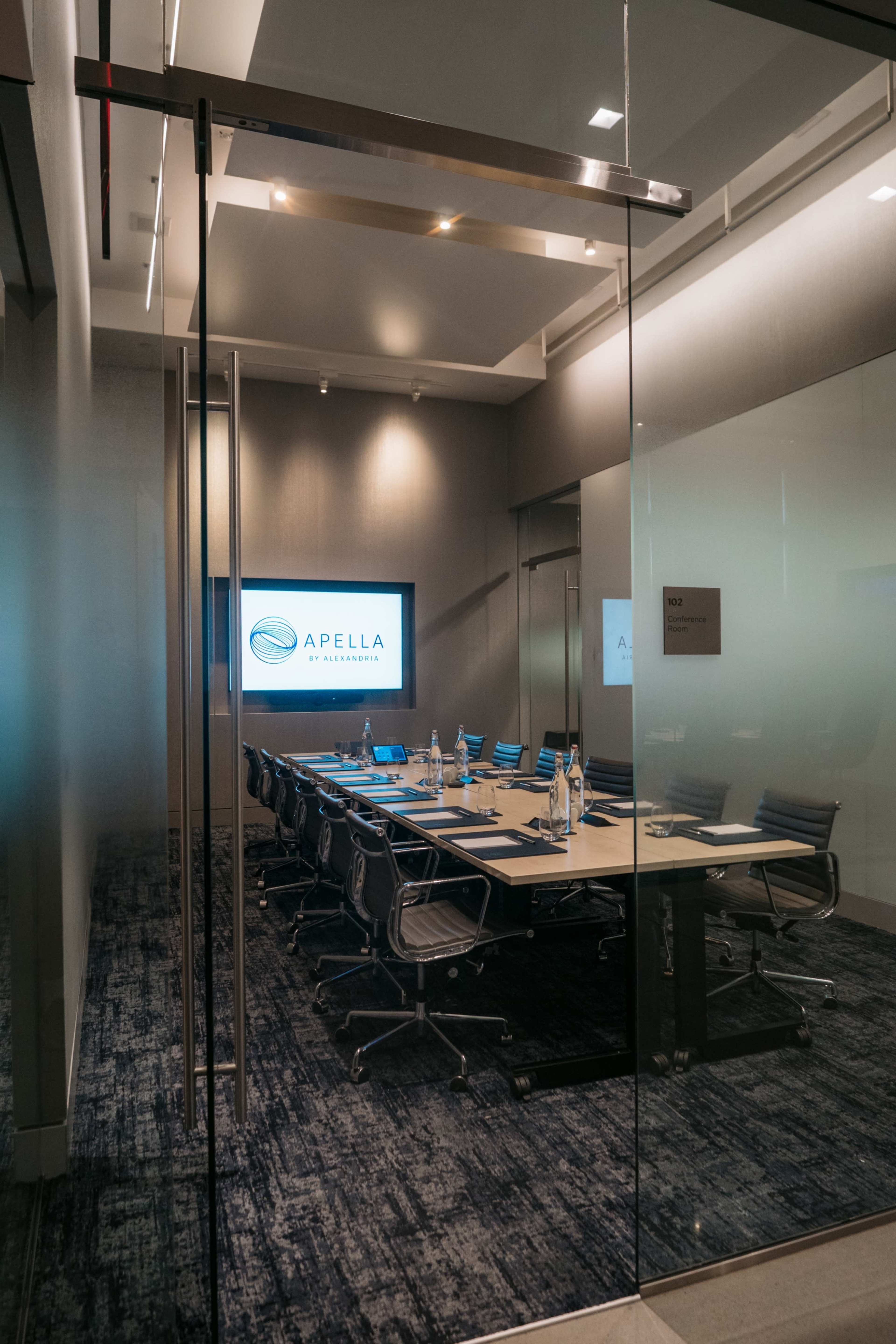A modern conference room with a large table, office chairs, and a screen displaying the logo "APELLA" is visible through glass doors.