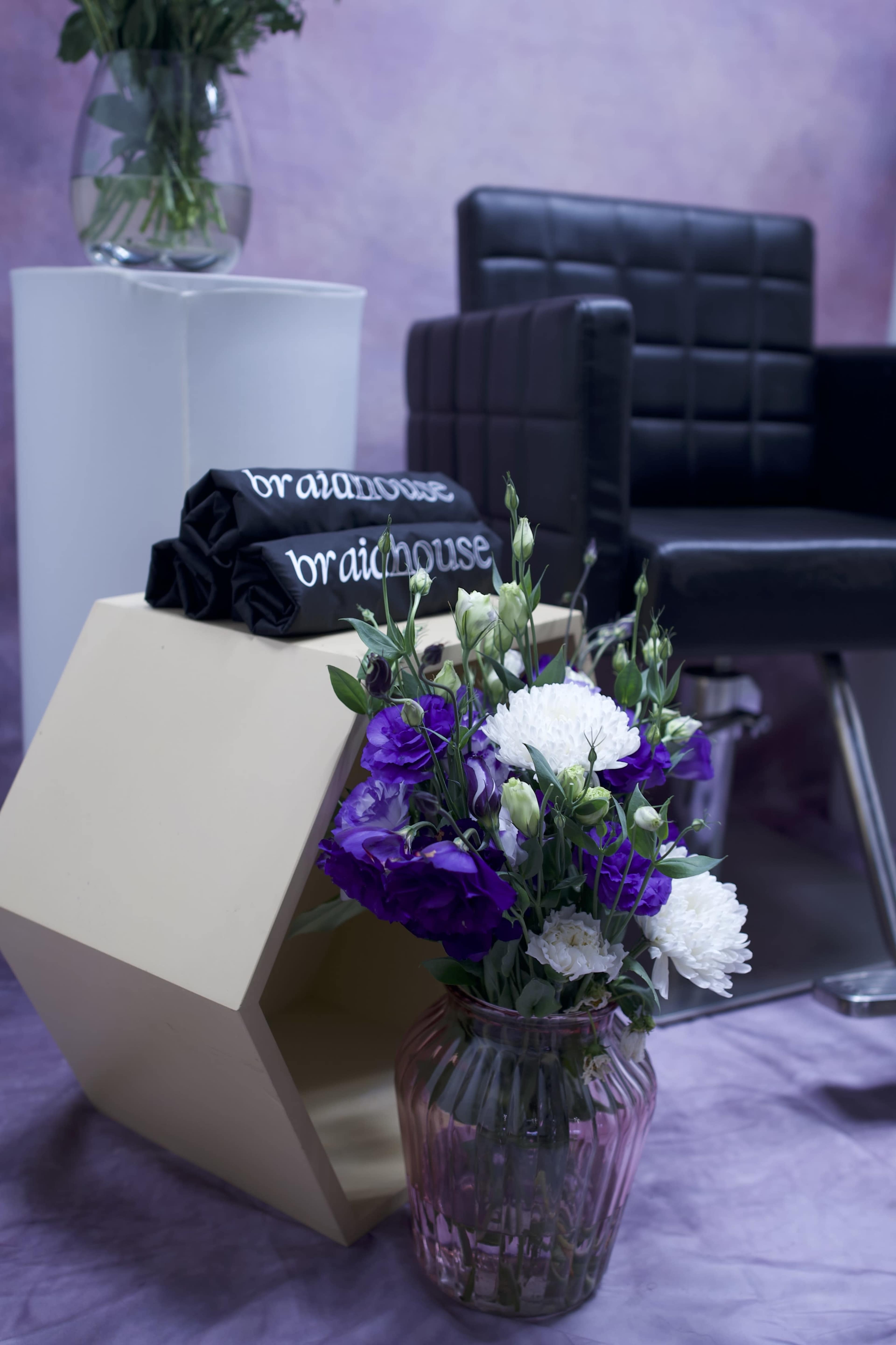 A black chair sits beside a hexagonal, beige storage unit topped with rolled black towels, next to a vase of purple and white flowers.