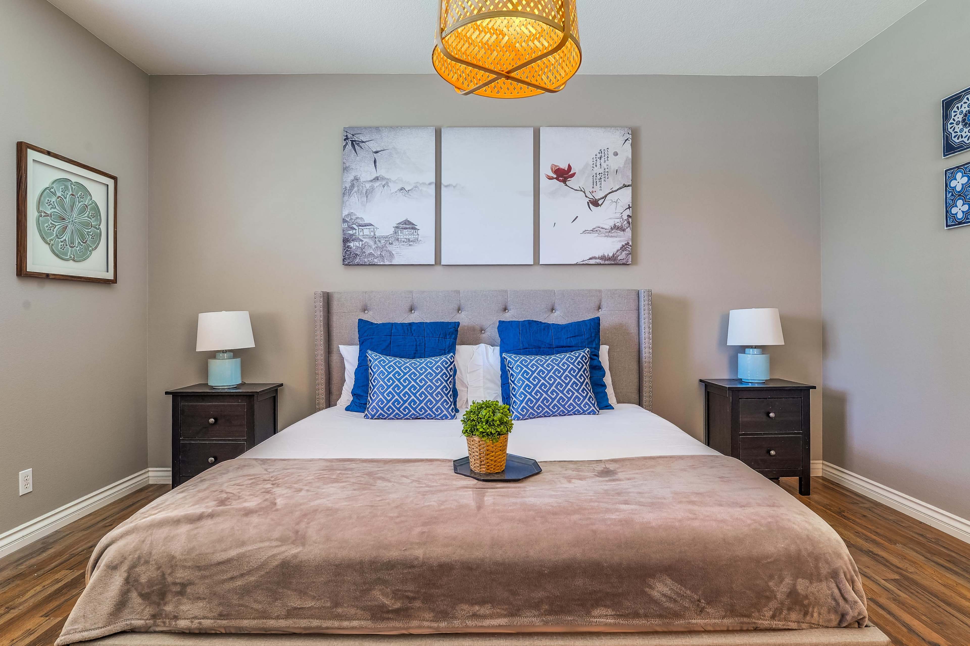 A neatly arranged bedroom with a bed featuring blue and white pillows, a wooden headboard, two bedside tables with lamps, and framed artwork on the walls.