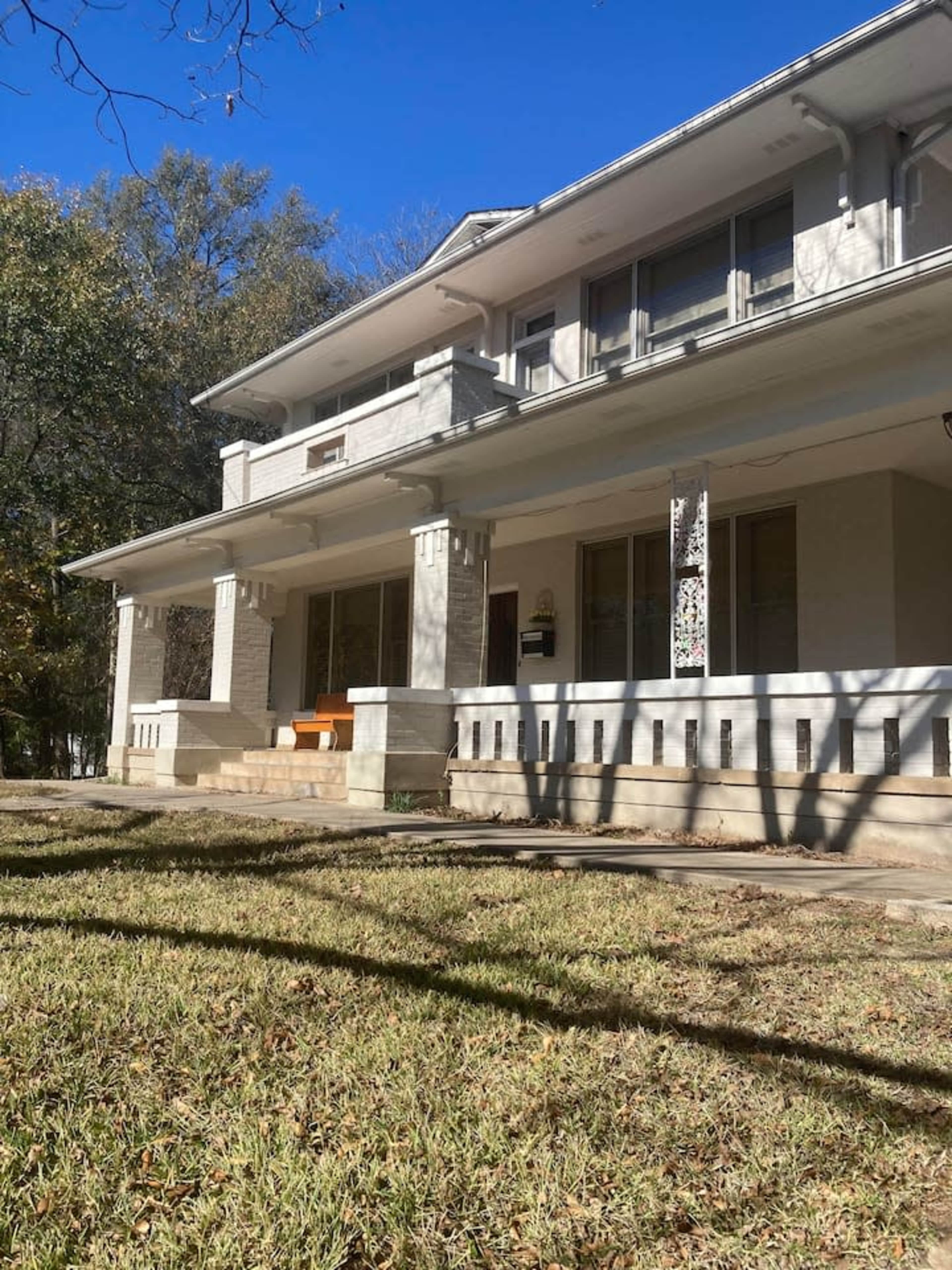 A two-story white house with a front porch and large windows surrounded by greenery.