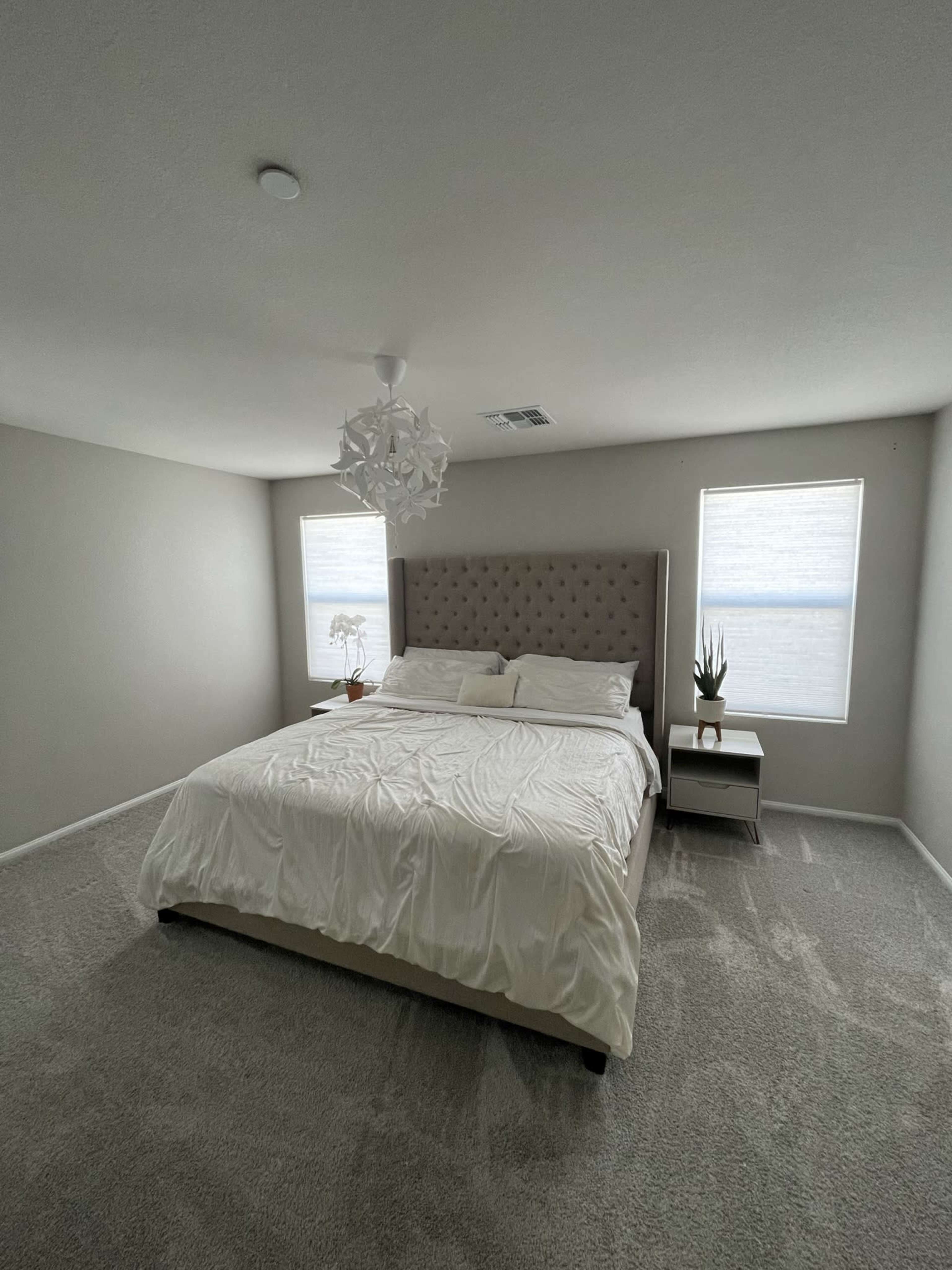 The image shows a spacious bedroom with a large bed, side tables, and two windows covered with light-colored shades.
