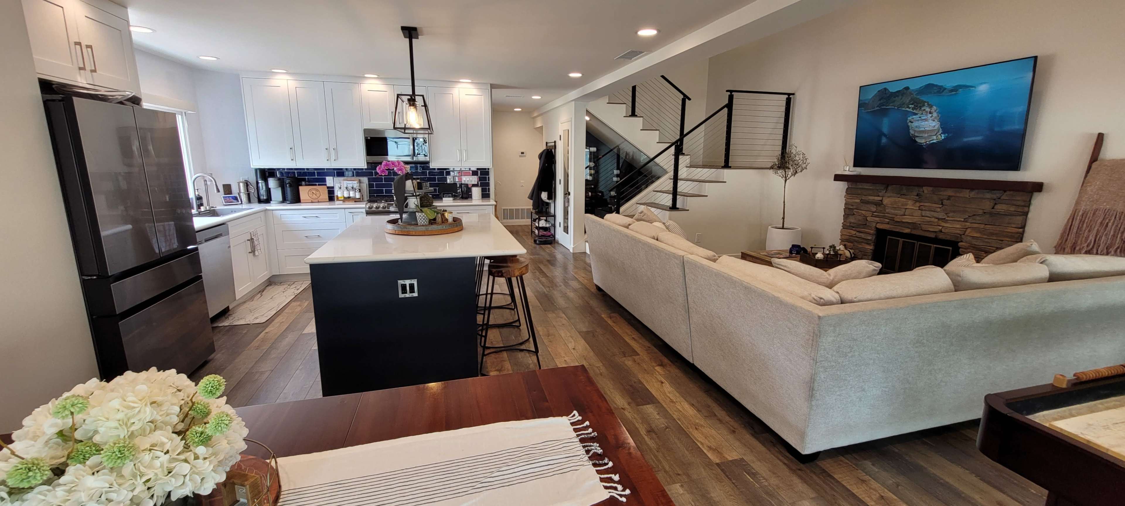 A modern open-concept living space featuring a kitchen with a large island, a dining area, and a cozy seating area with a view of a staircase and a stone fireplace.