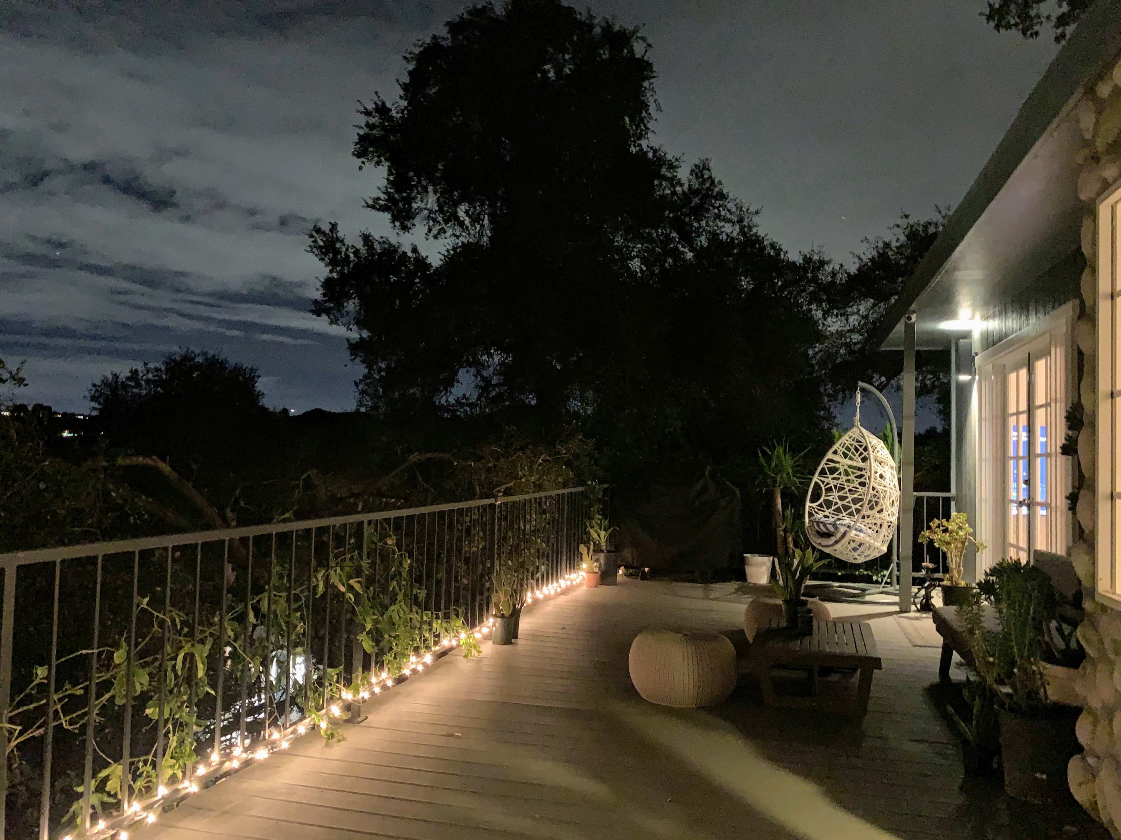 A wooden deck at night, illuminated by string lights, featuring a hanging chair and potted plants.