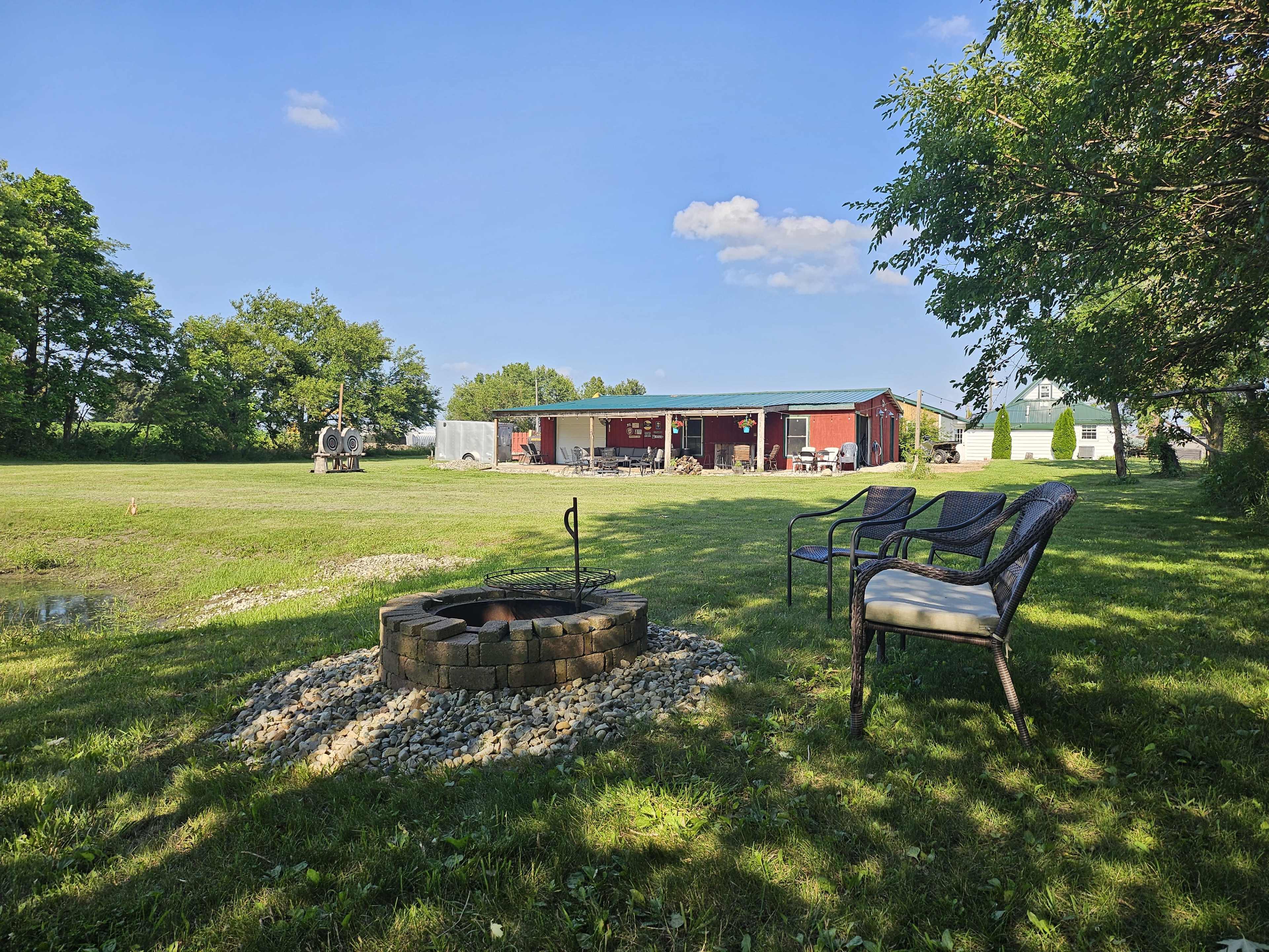 A fire pit made of stone is surrounded by chairs in a grassy area with a red barn and fields in the background under a clear blue sky.