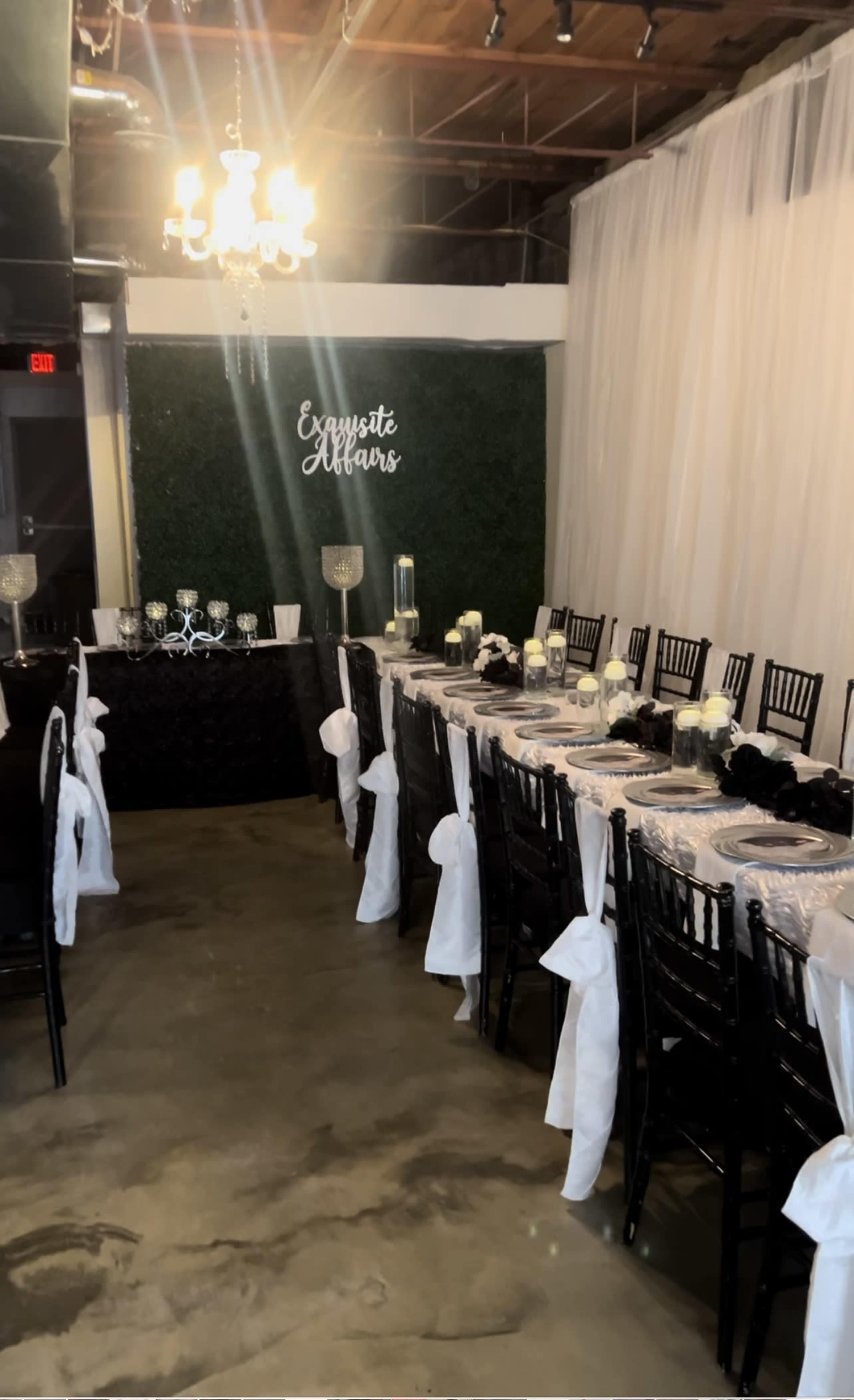 A long dining table adorned with white tablecloths and black accents is set up in a banquet space featuring greenery on the wall and a chandelier overhead.