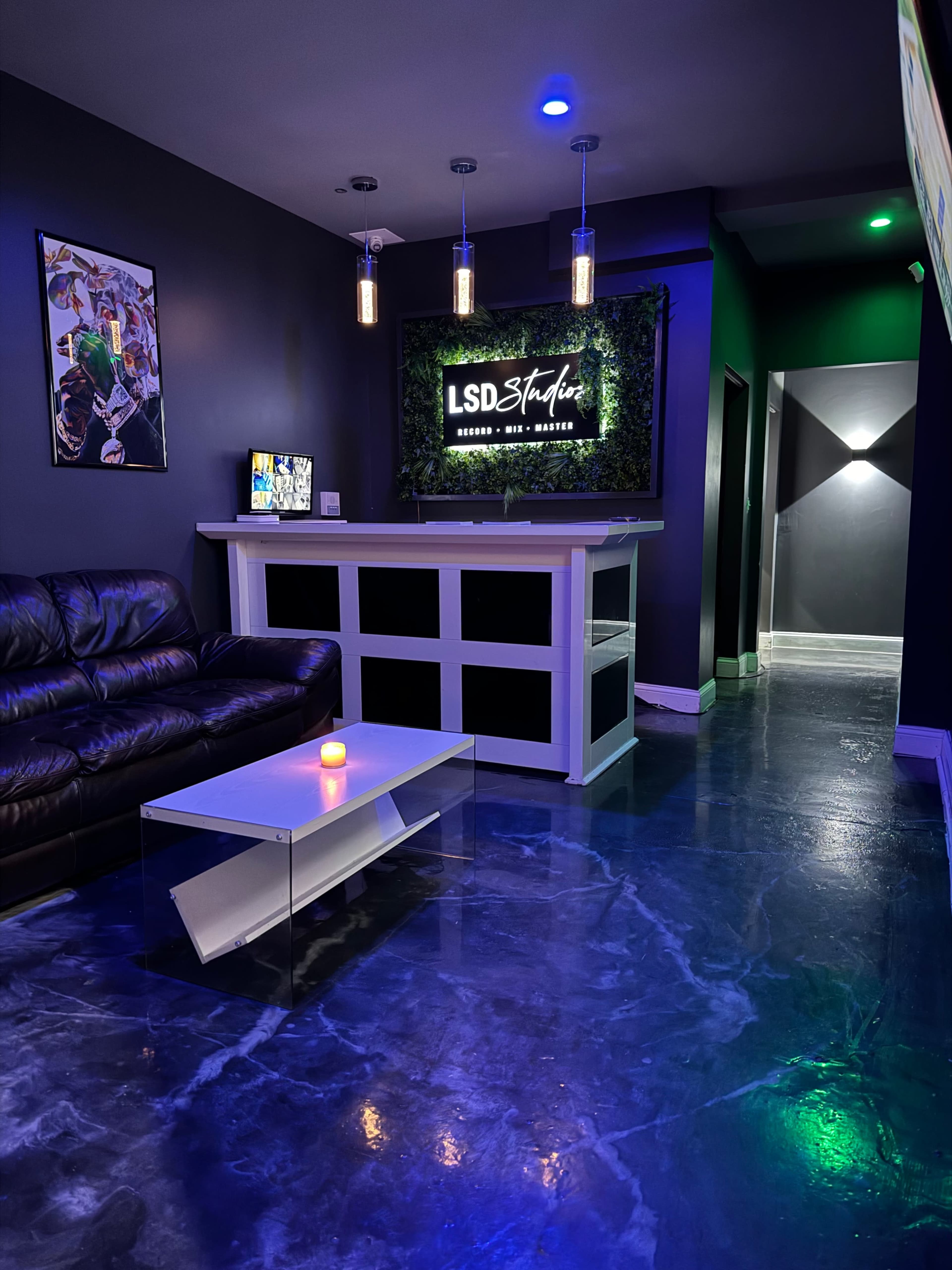 The image shows a modern reception area with a dark-painted wall, a sleek white counter with a sign that reads "LSD Studios," and a leather couch accompanied by a coffee table, illuminated by pendant lights.