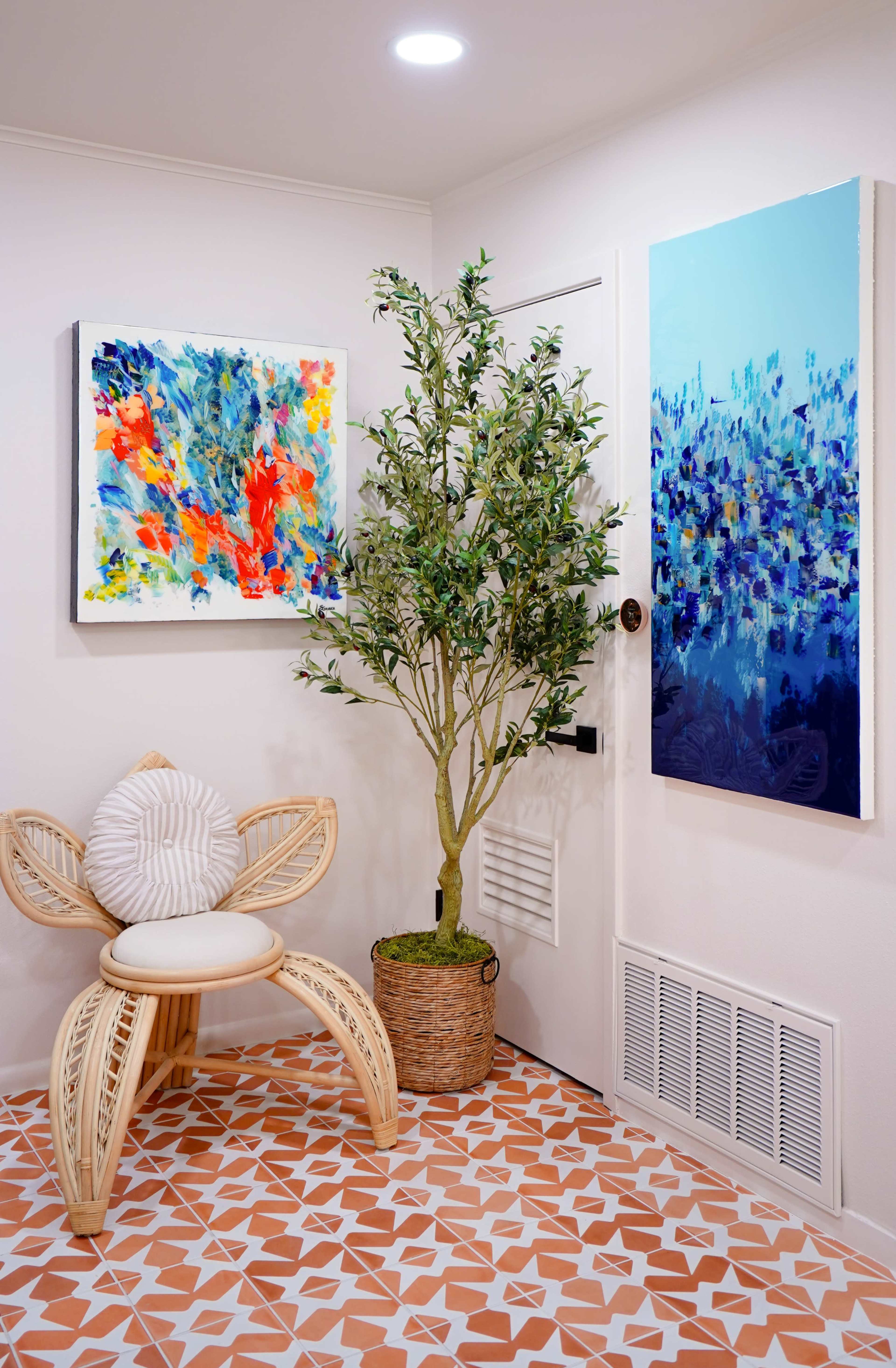 A plant stands next to a decorative chair in a corner with two colorful paintings hanging on the walls and a patterned tile floor.