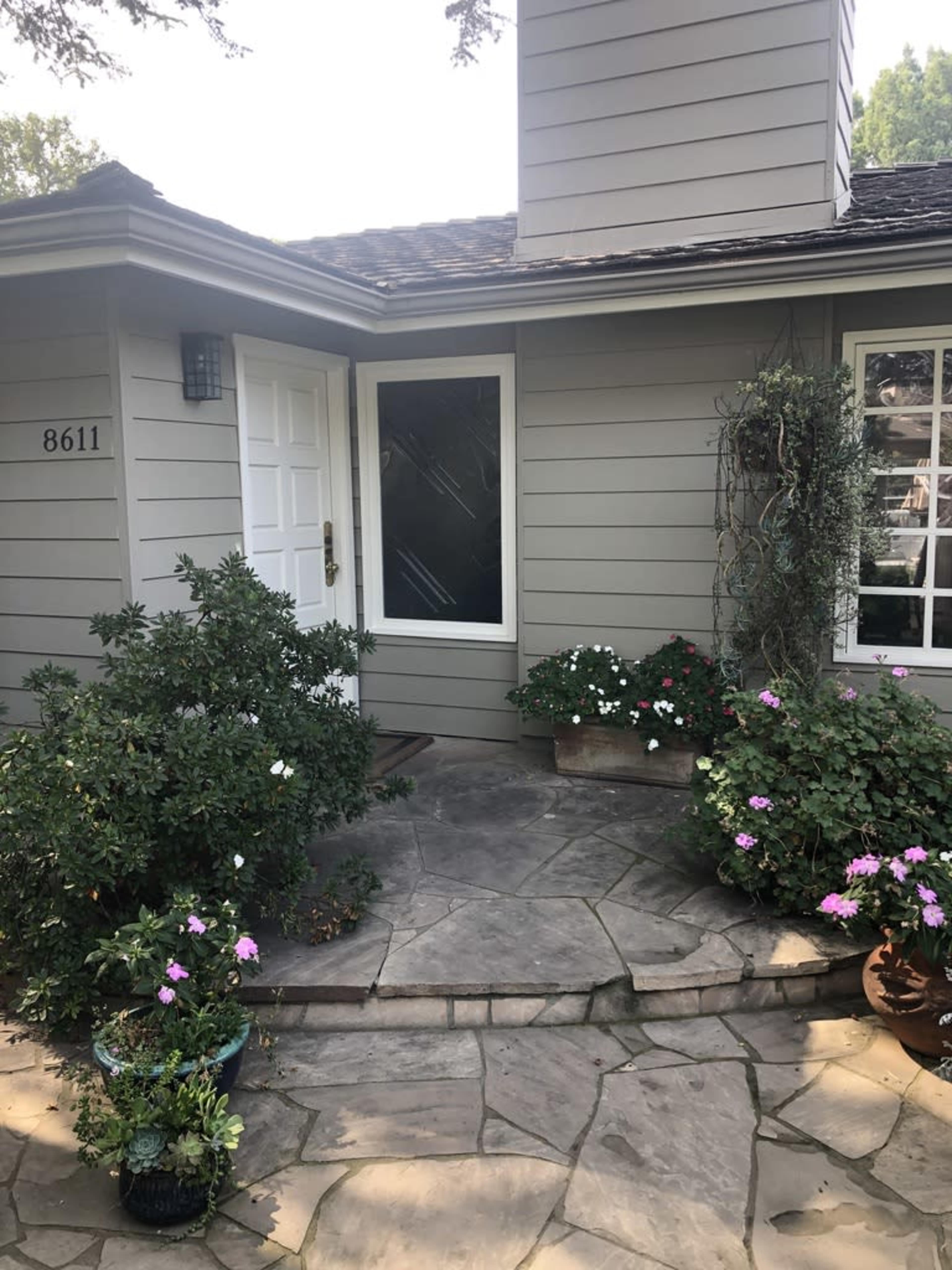 A house entrance with a stone walkway, surrounded by potted plants and flowers near the door.
