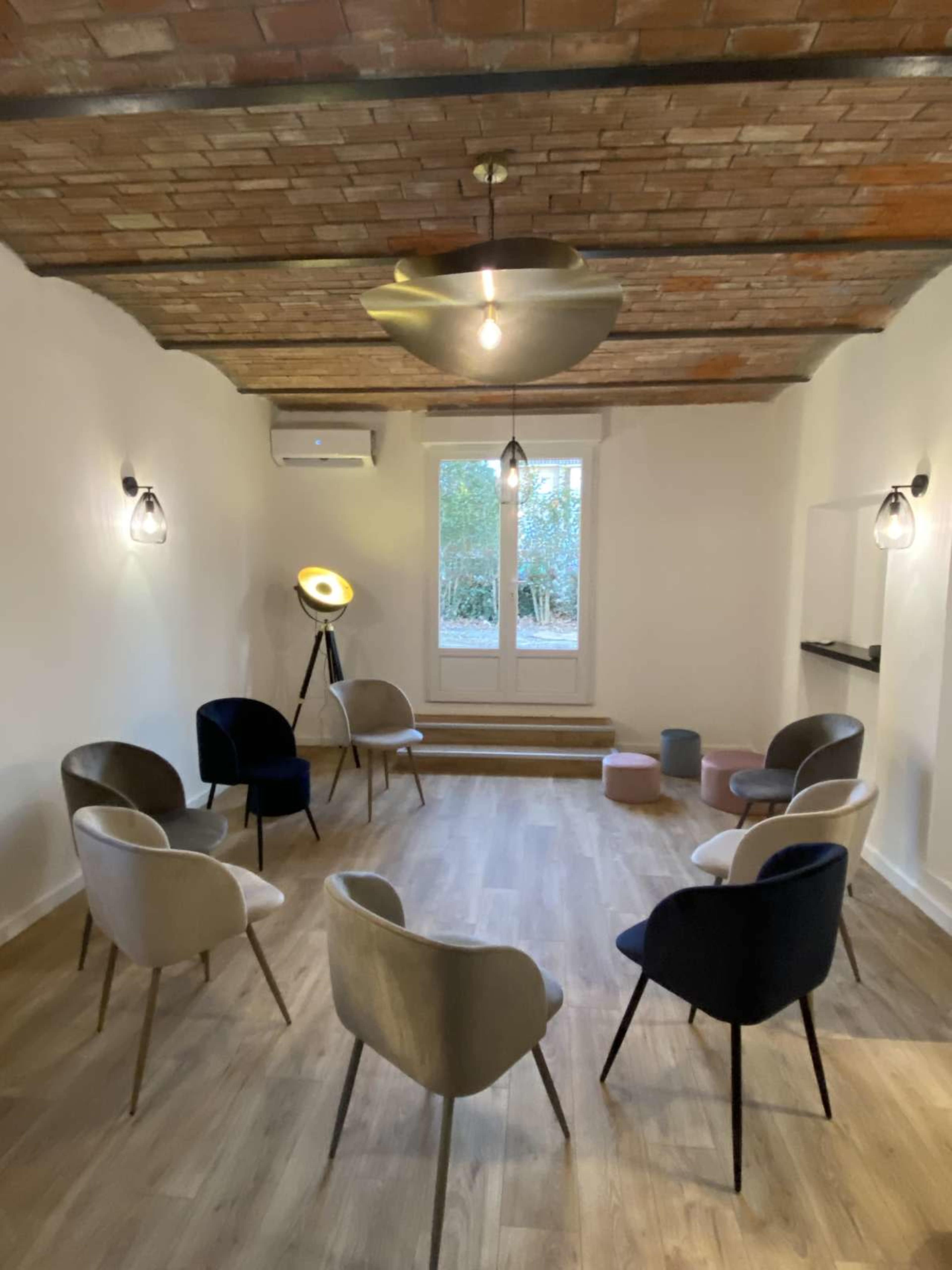 The image shows a minimalist meeting room with a circular arrangement of chairs, a tripod light, and a large window looking out into a green area.