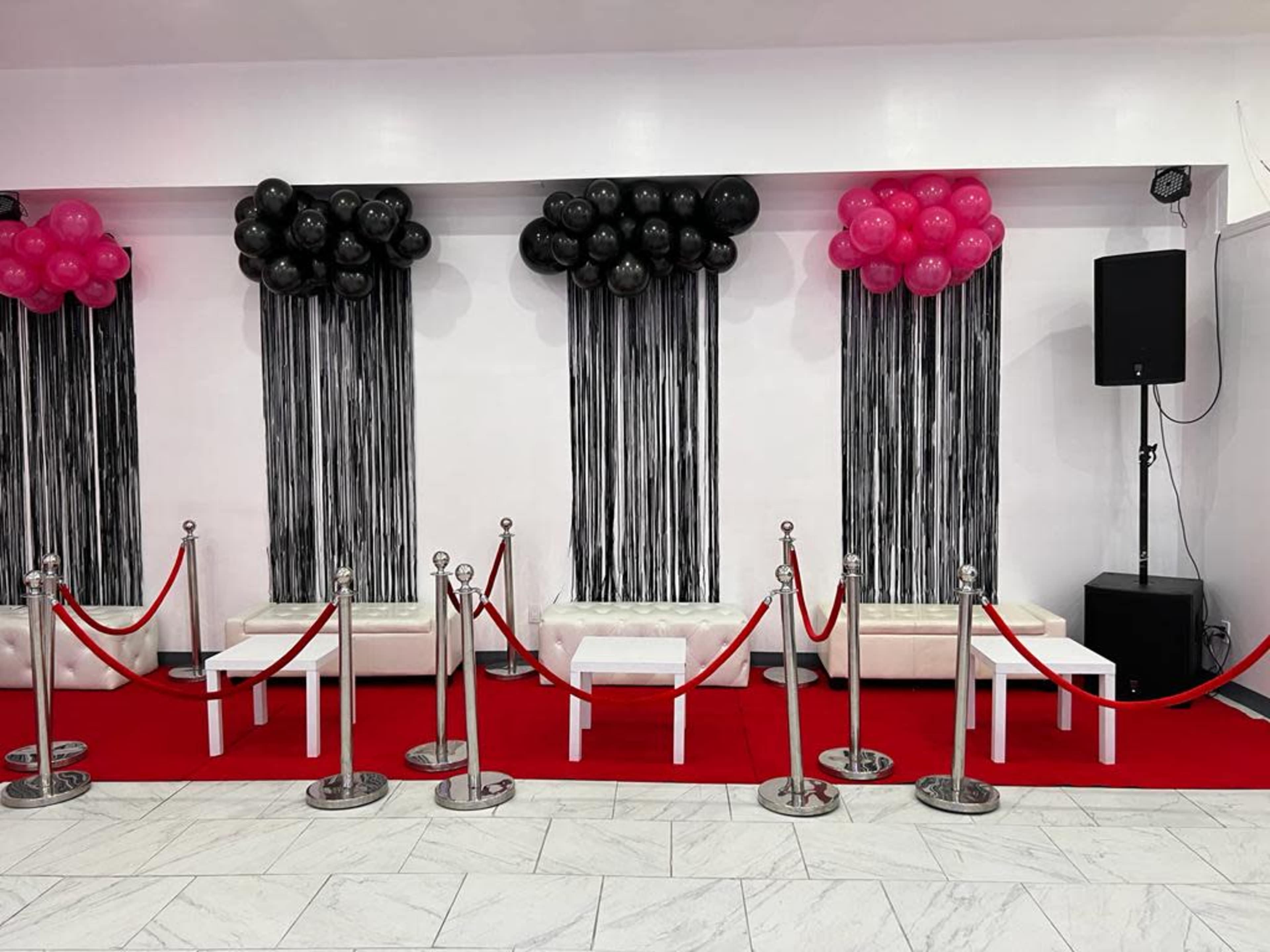 A section of an event space features black and pink balloon decorations, red carpet, and a velvet rope barrier with metallic stanchions.