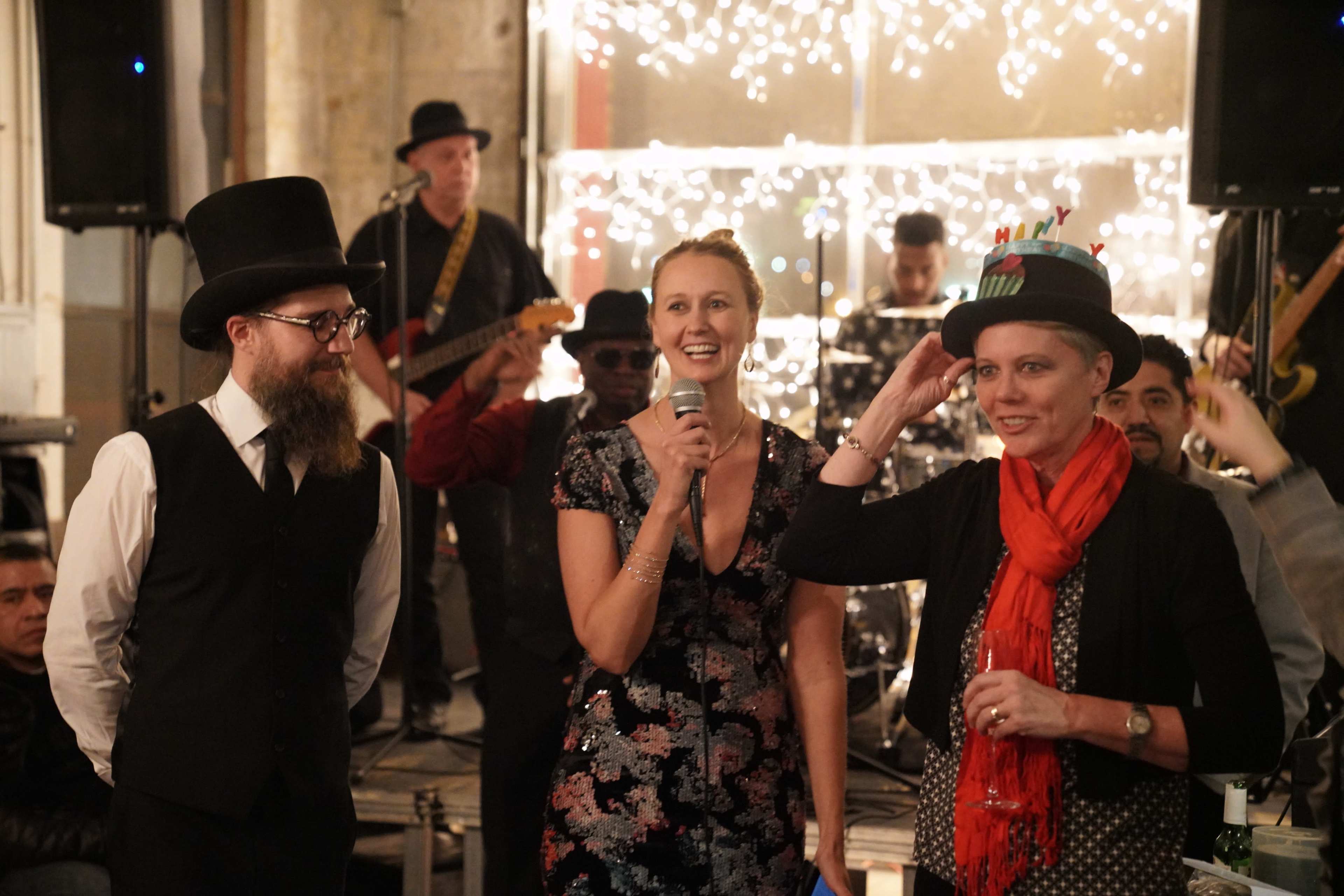 A woman speaks into a microphone while two people, one in a top hat and the other in a festive hat, stand beside her in front of a band performing on stage.
