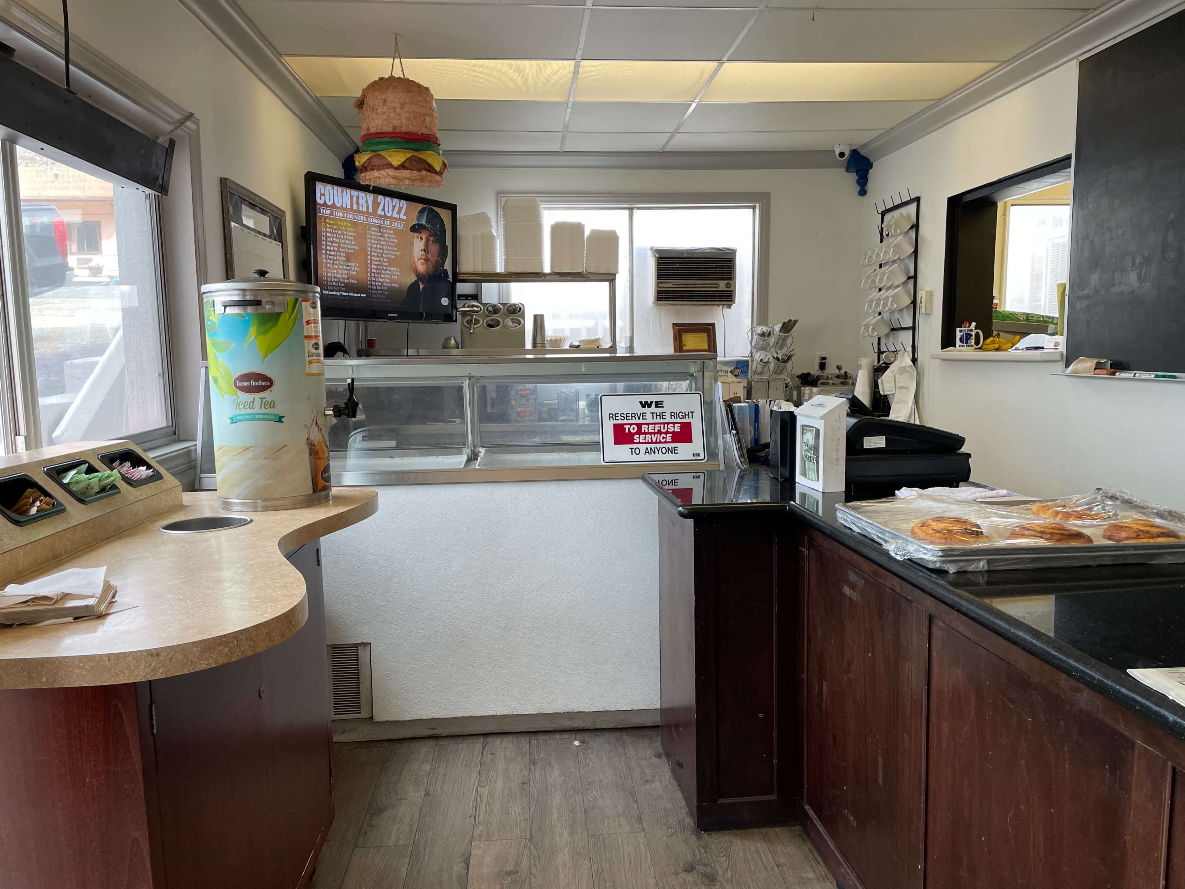 A small restaurant interior features a counter with menu displays, a cash register, and various food items visible on the shelves.