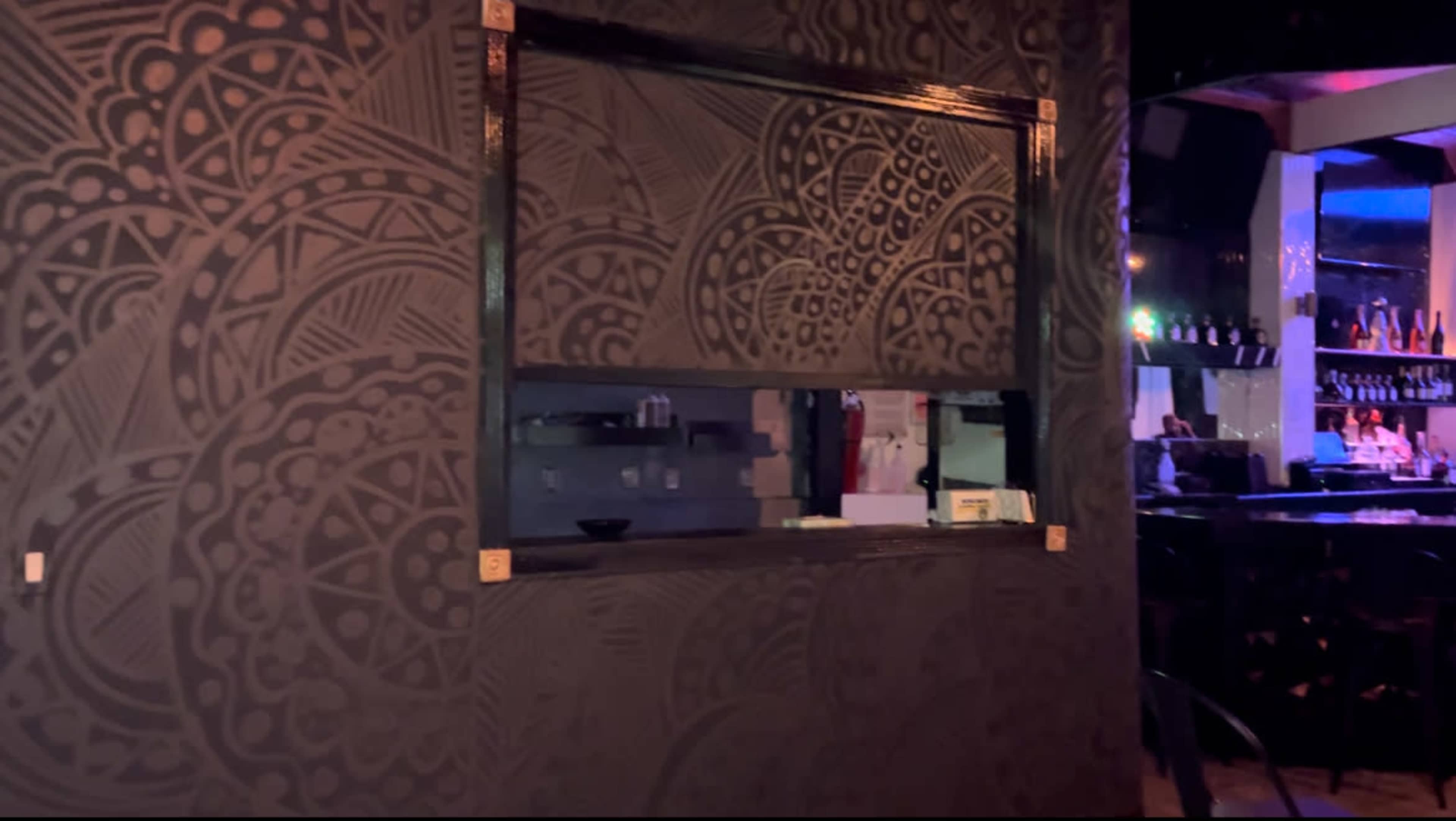 The image shows a darkly patterned wall with an opening that leads to a bar area, featuring shelves with liquor bottles illuminated by colored lights.