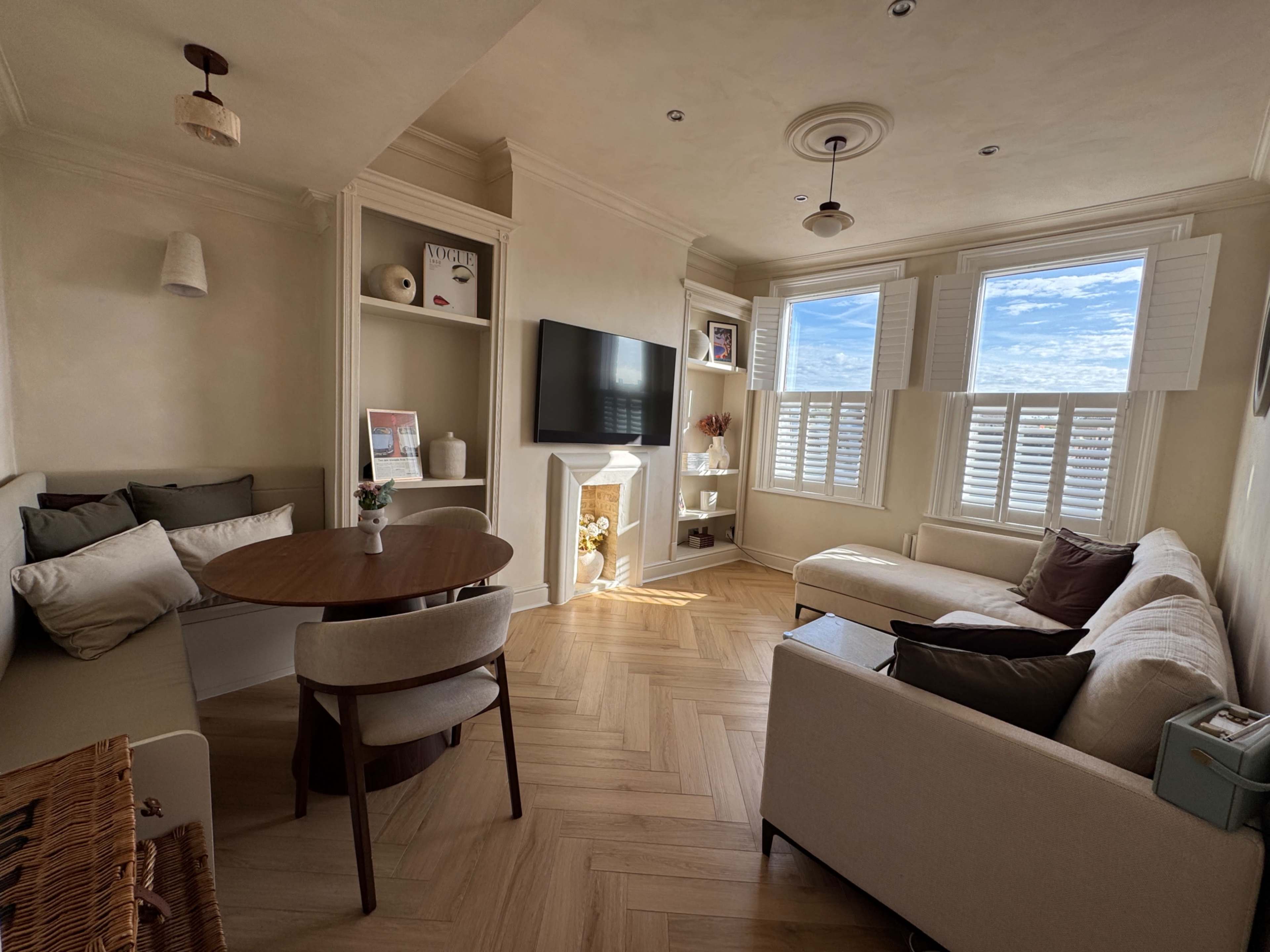 The image shows a cozy living room with a round dining table, a sofa, a television mounted on the wall, and large windows letting in natural light.