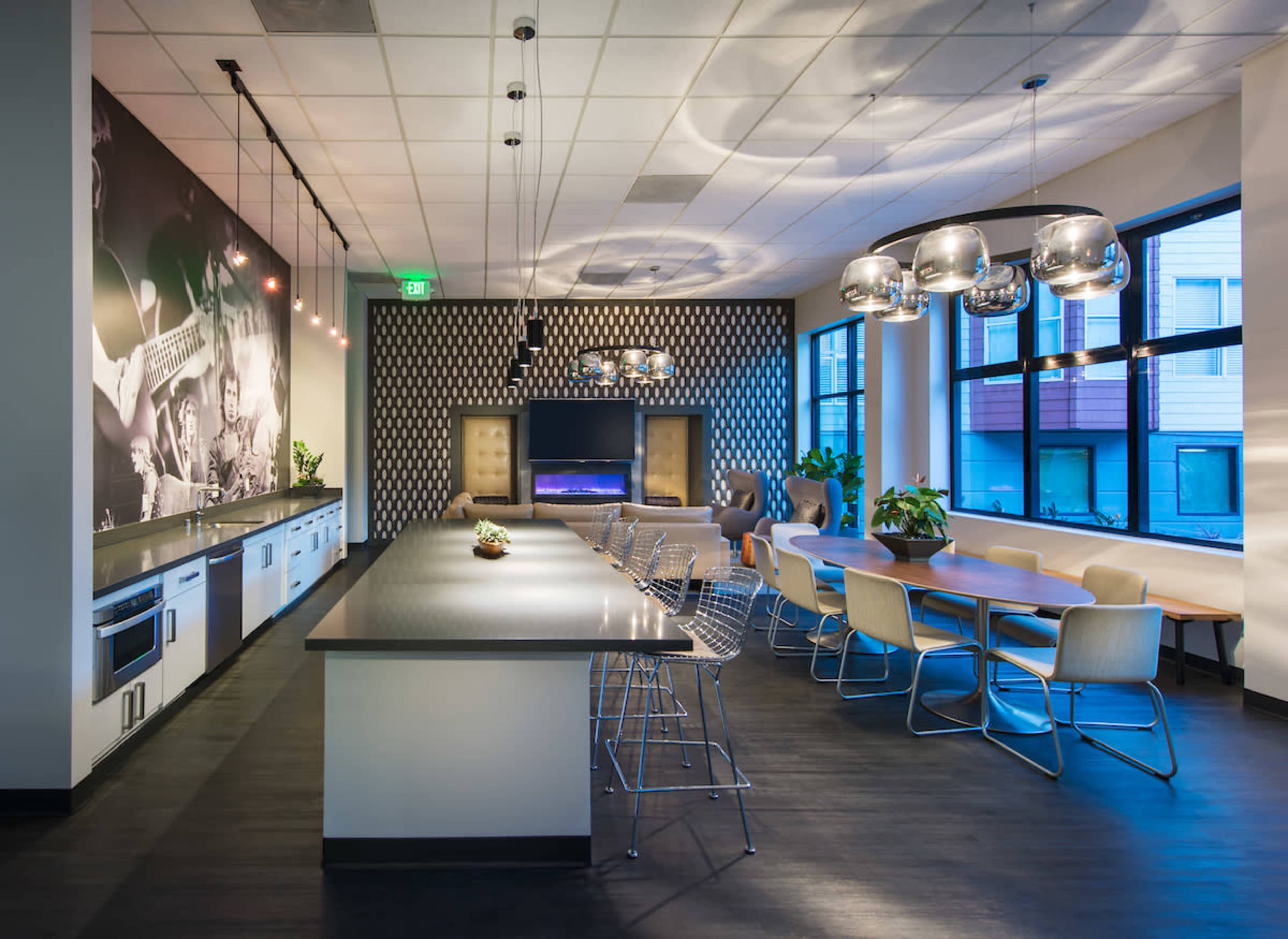 A modern communal kitchen and dining area with a long countertop, chairs, and a large television on a patterned wall.