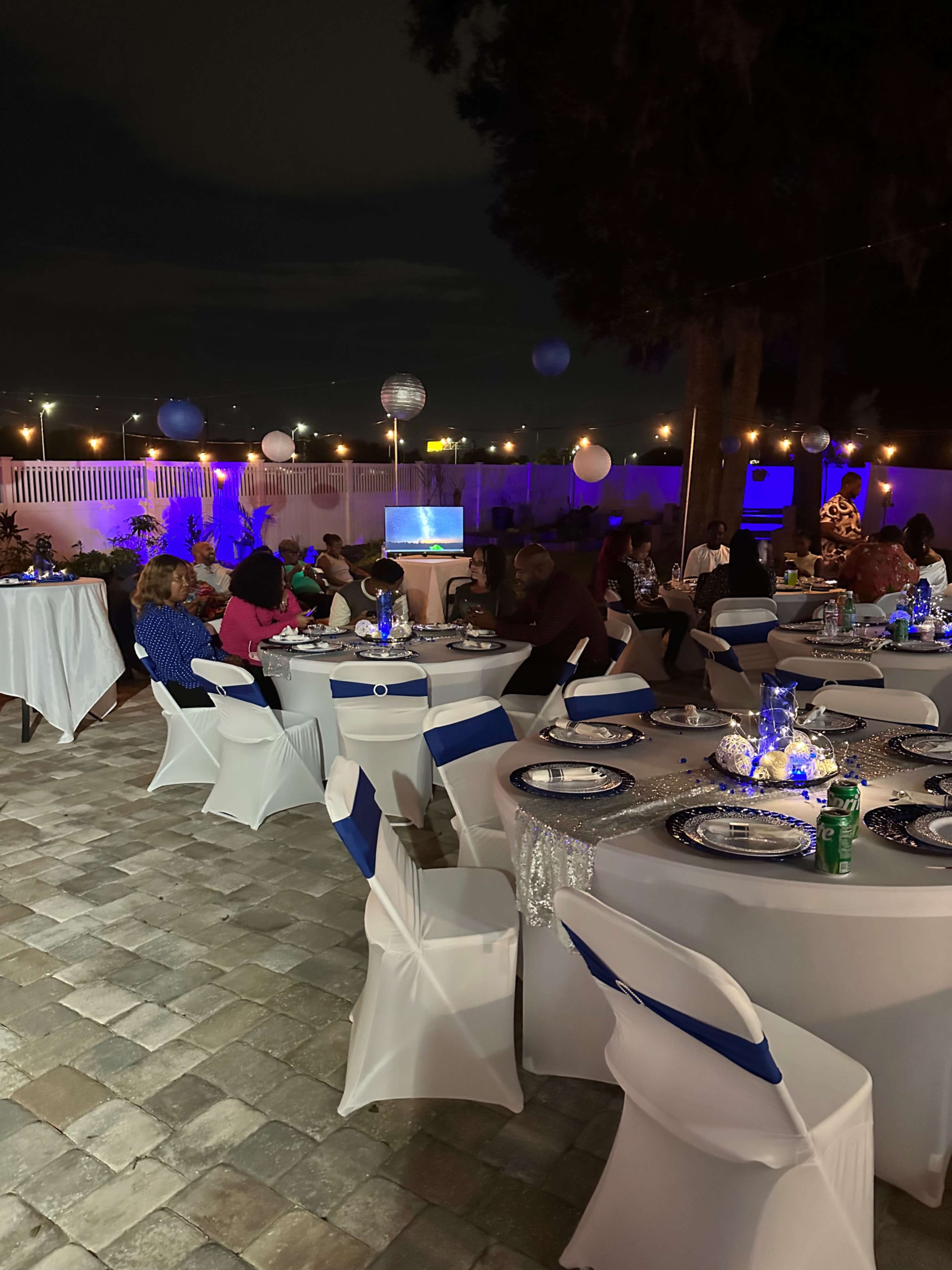 A decorated outdoor event space features tables with white tablecloths and blue accents, filled with guests enjoying the evening under soft lighting.