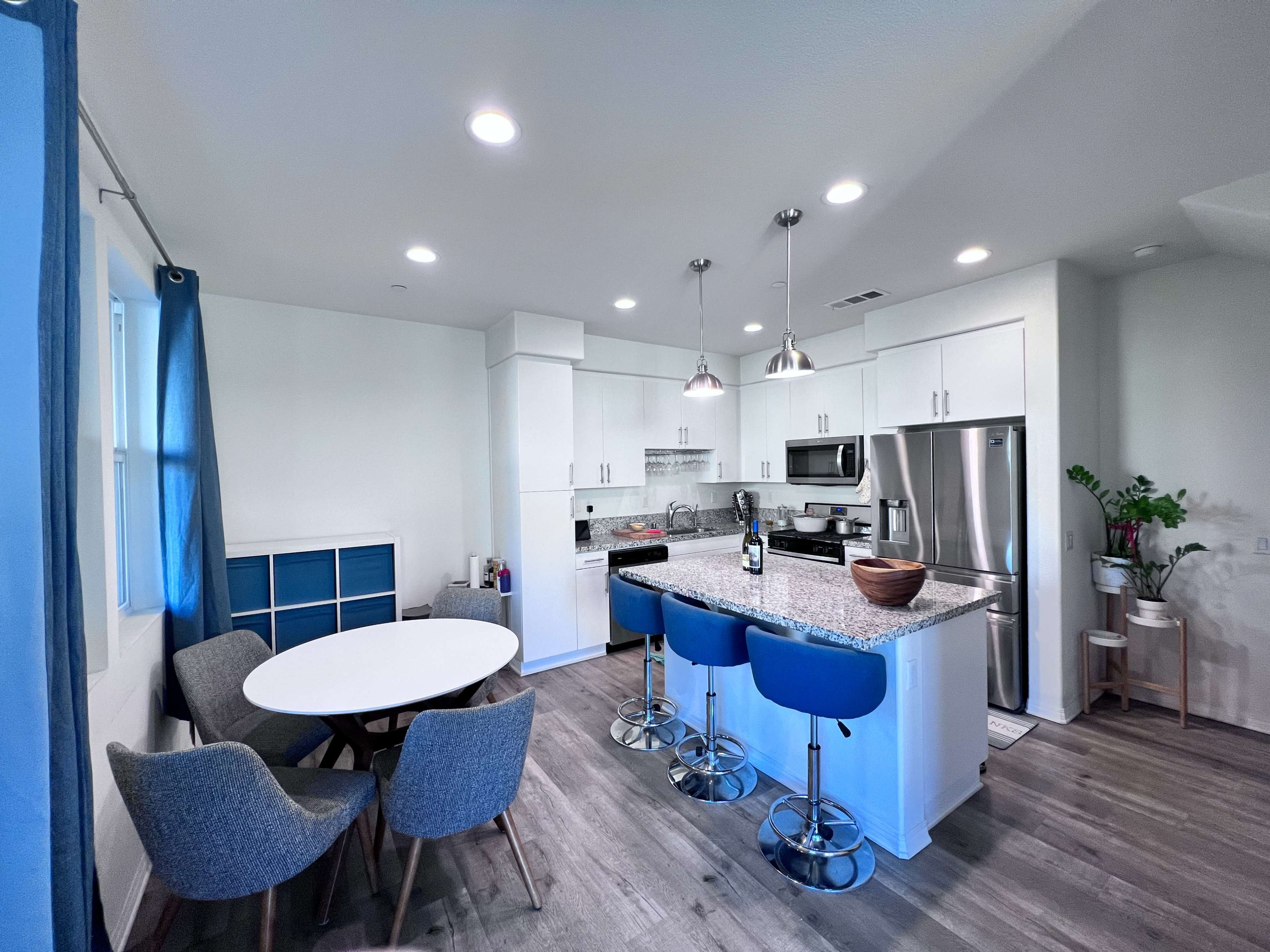 A modern kitchen features white cabinets, a gray countertop, stainless steel appliances, and a dining area with a round table and blue chairs.