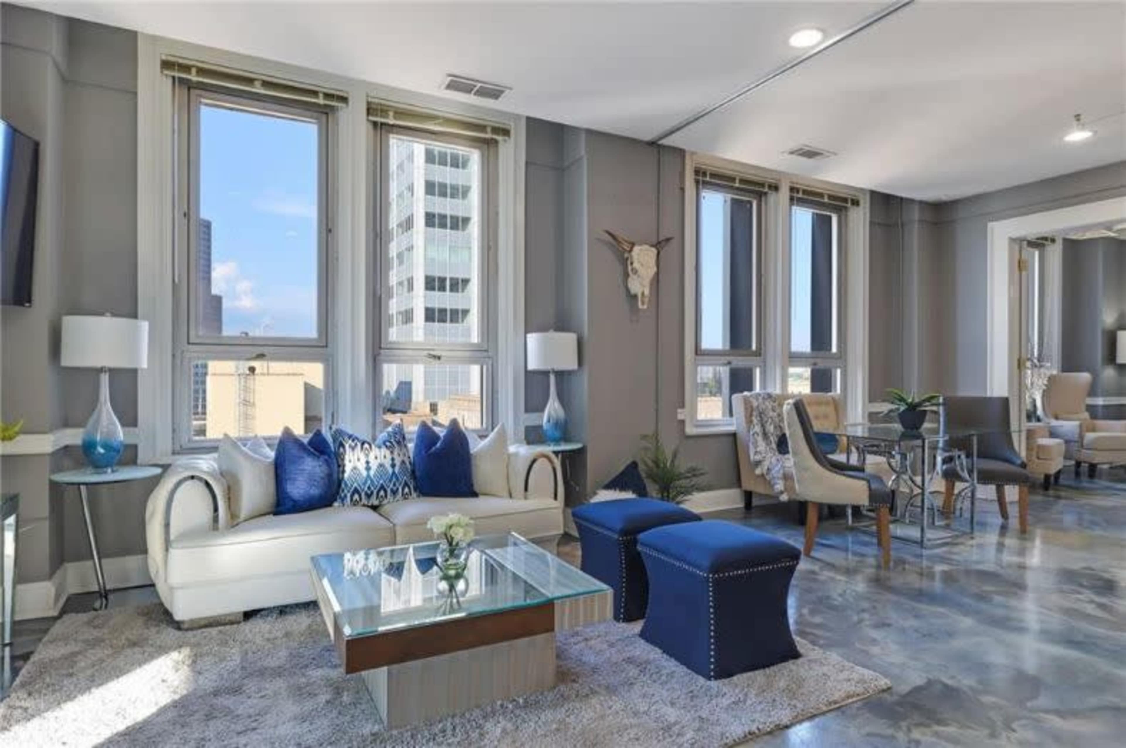 A modern living room features a cream-colored sofa with blue accent pillows, a glass coffee table, and large windows offering a view of city buildings.