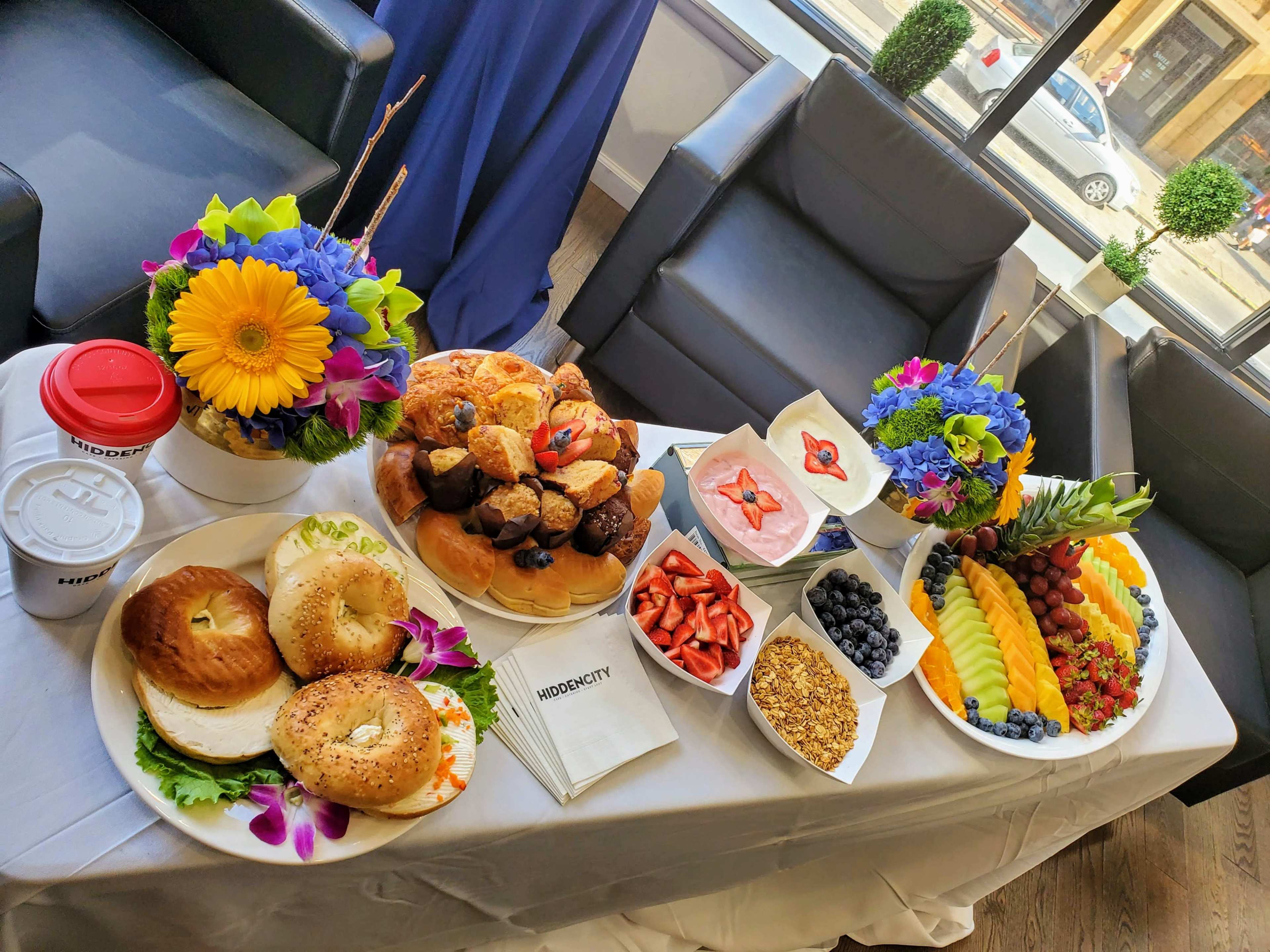 A diverse spread of breakfast foods, including bagels, pastries, fruits, and coffee, is arranged on a table next to comfortable seating.