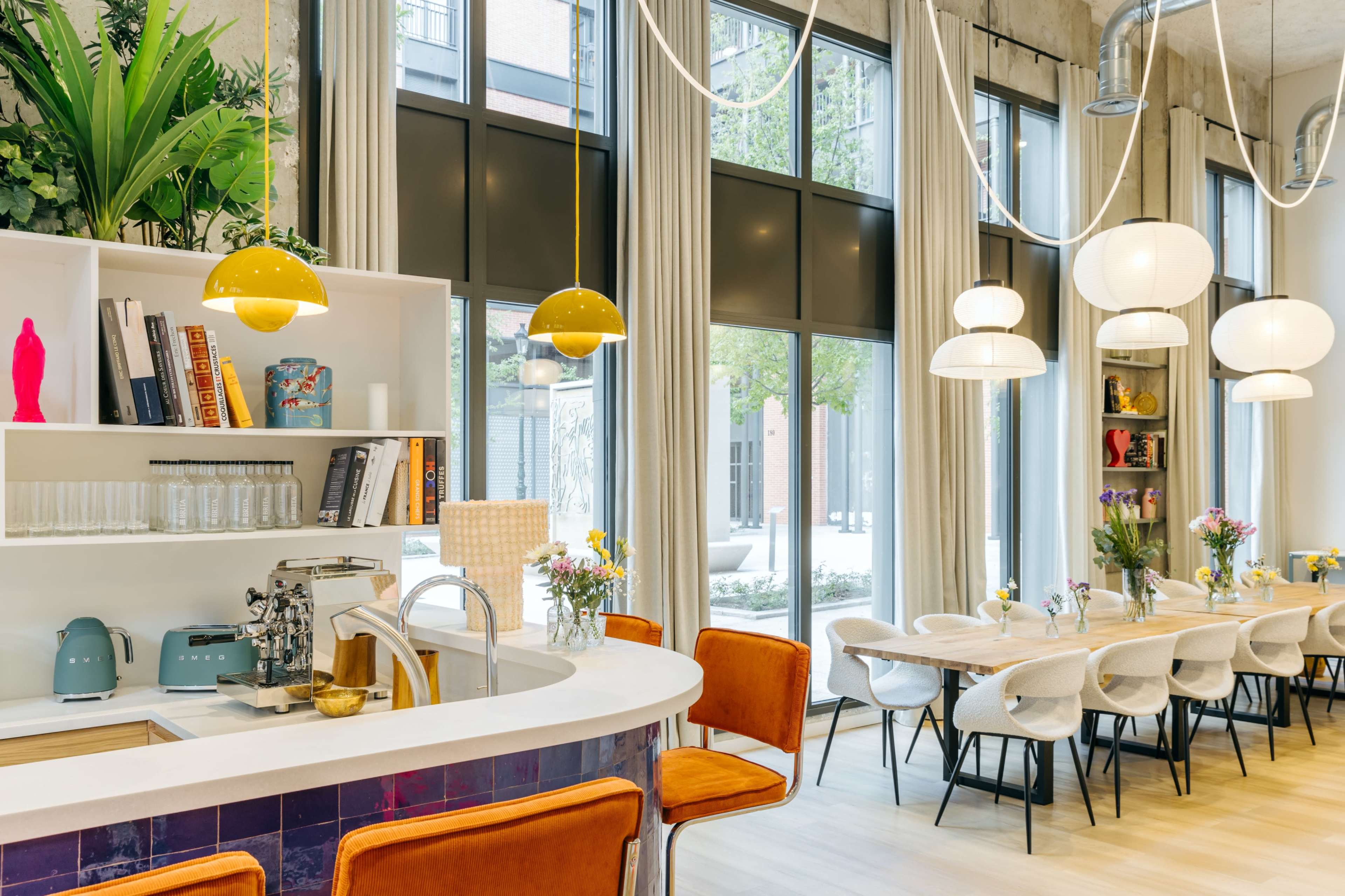 A bright and modern café features a white countertop bar area with teal appliances, surrounded by large windows and a long dining table set for guests.